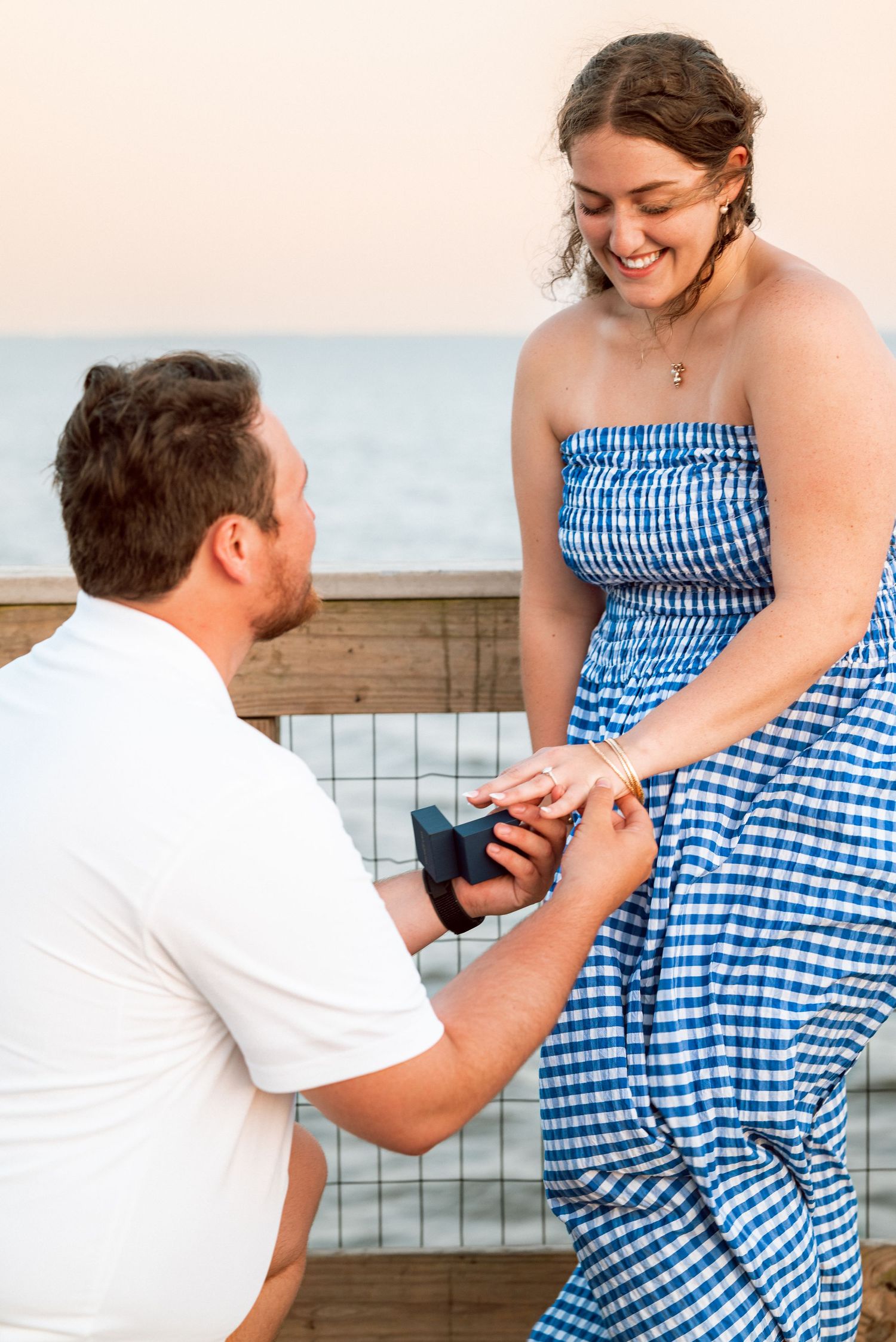 Beach proposal moment with blue gingham dress and engagement ring box.