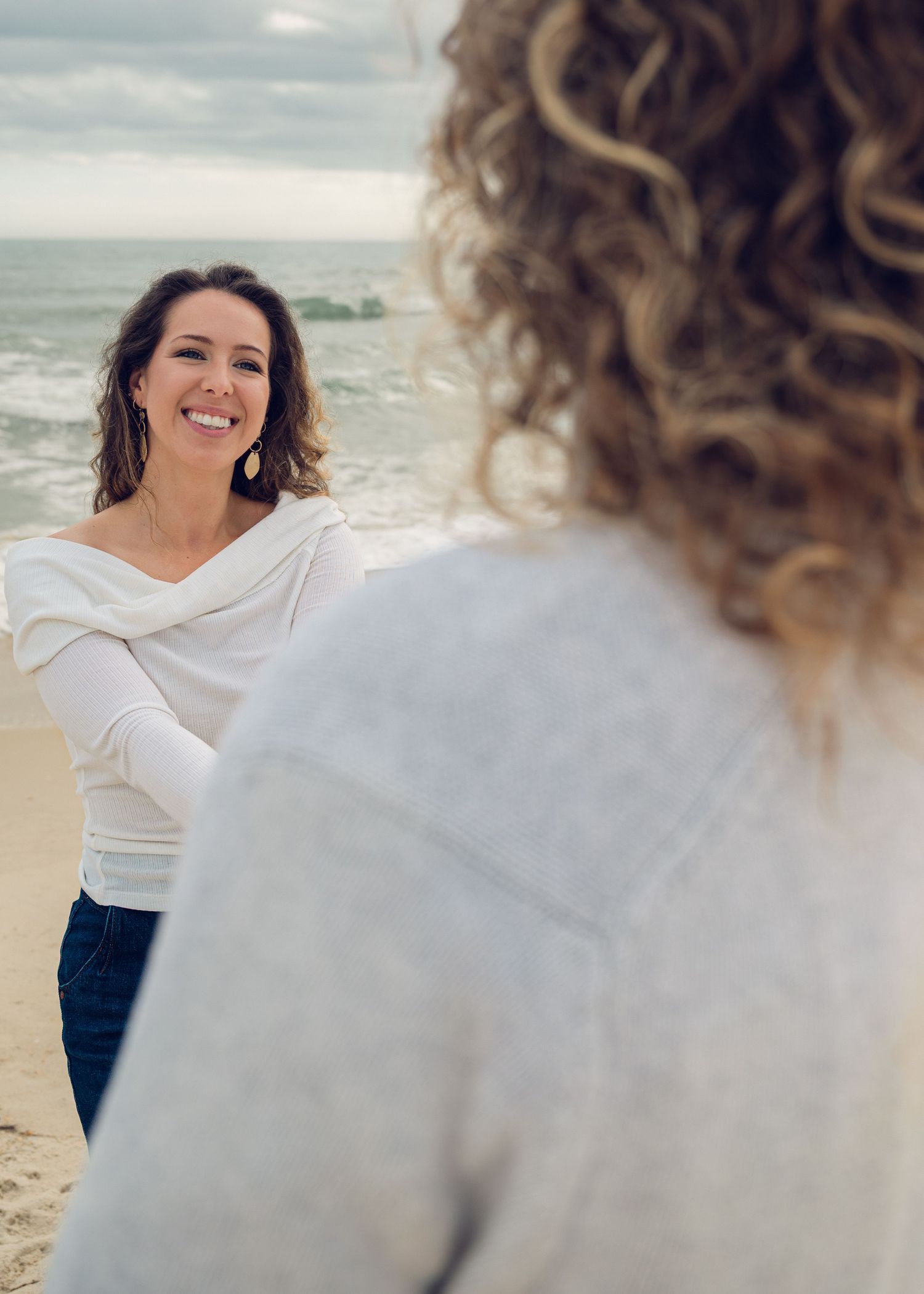 Two people in white tops having a candid conversation on a peaceful beach with ocean waves in the background.
