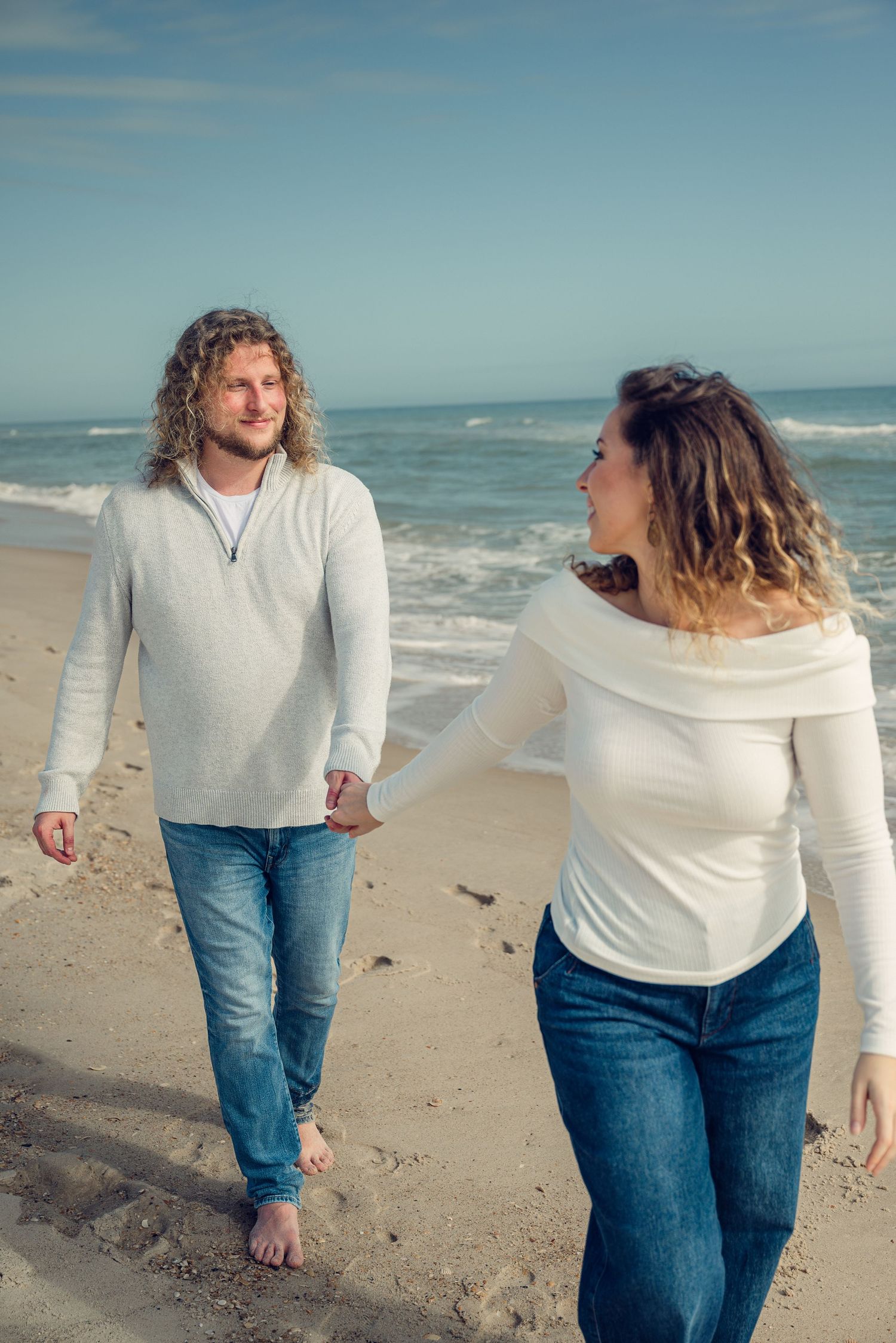 Two people in white sweaters and jeans walking barefoot along a beach with ocean waves in the background.