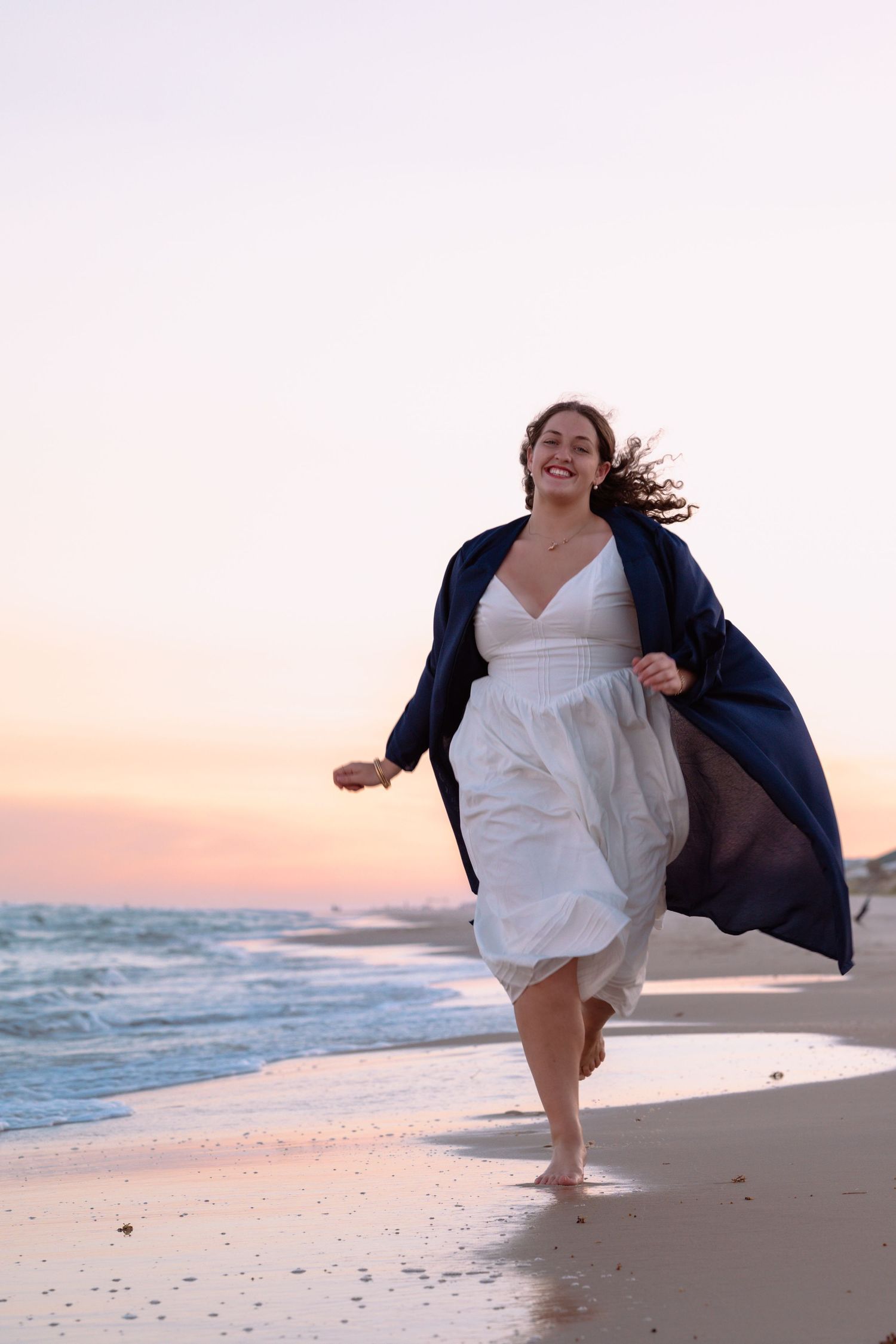 A graduate in a white dress runs joyfully along the shoreline during sunset.