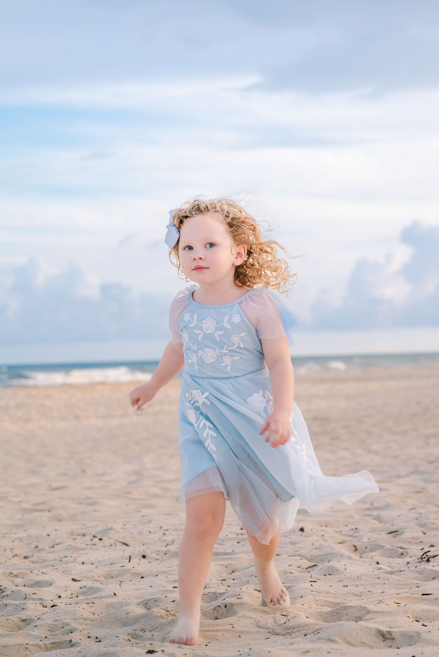 A curly-haired toddler girl in a light blue dress walks barefoot on a sandy beach with ocean waves in the background.