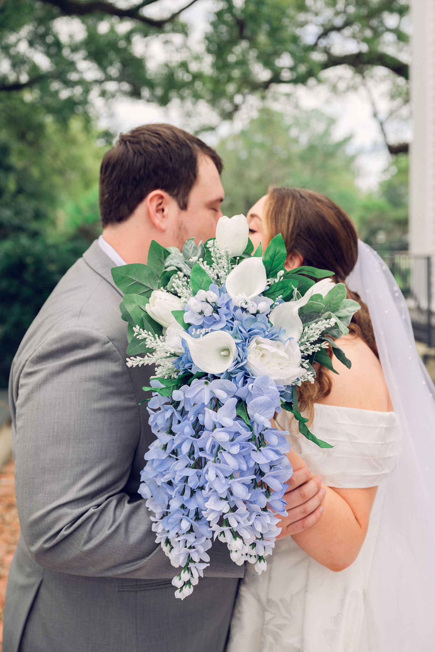 Bride and groom share a kiss behind a cascading bouquet of blue hydrangeas and white roses on their wedding day.