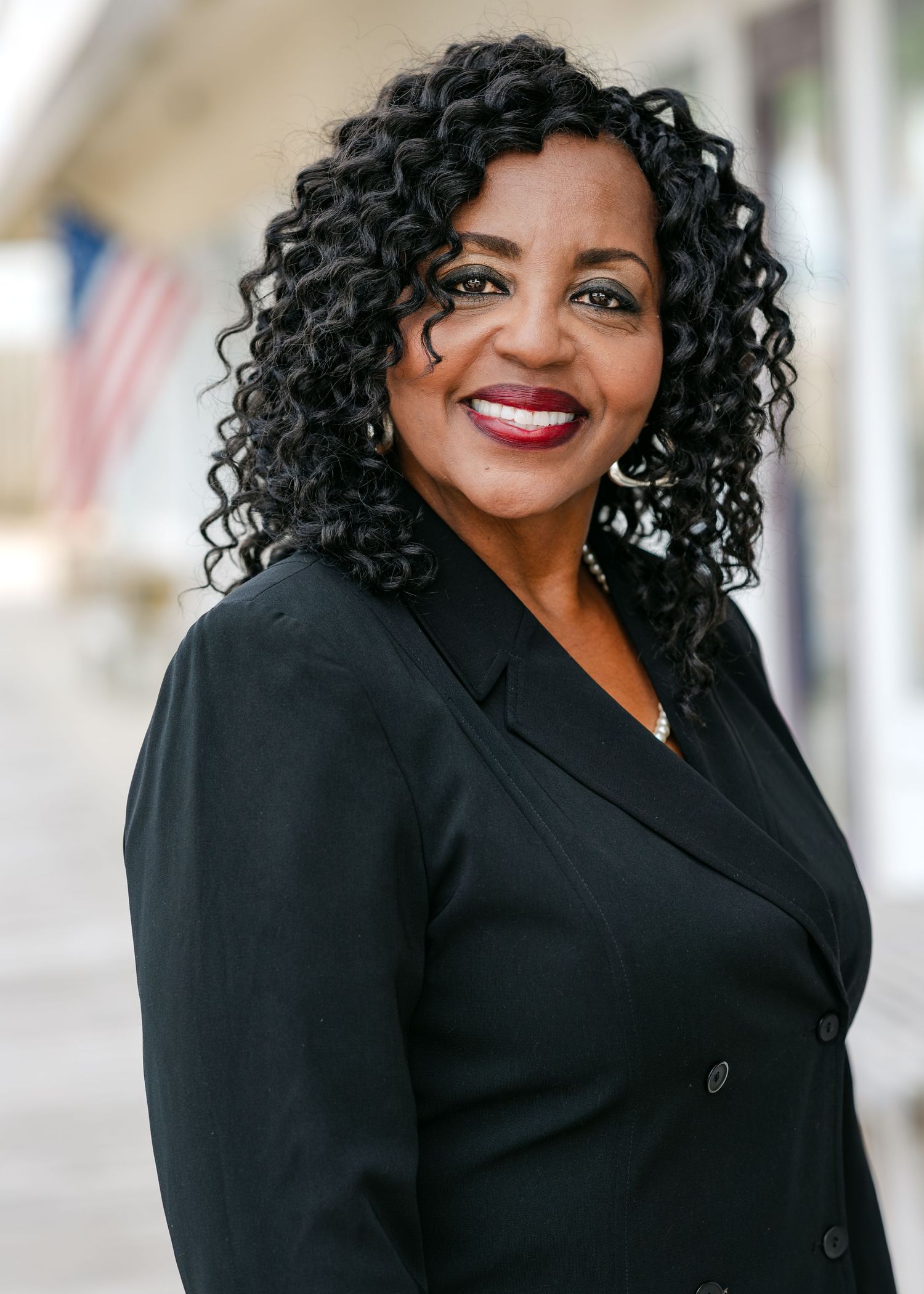 Professional woman in black blazer smiling confidently outdoors with an American flag in the background.