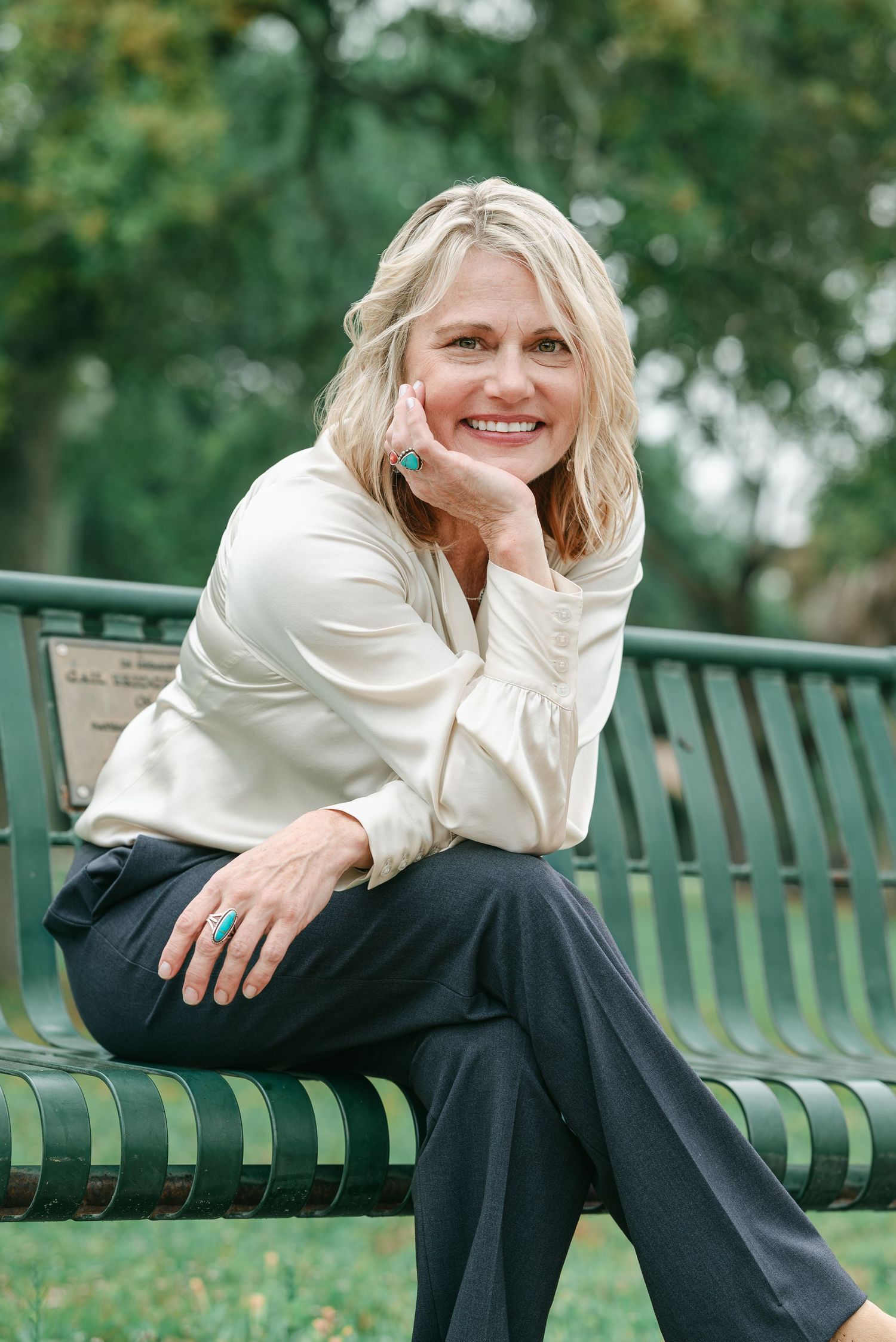 Smiling blonde woman in cream blouse and dark pants sitting on a green park bench outdoors.