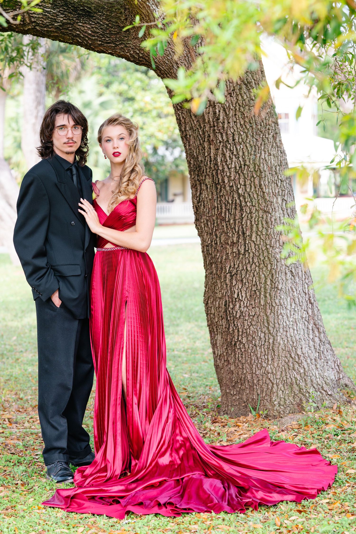 Couple in formal wear poses dramatically with flowing red dress train in autumn garden setting.