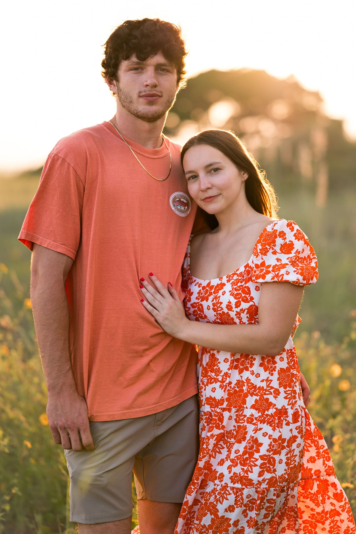 A couple in orange and floral attire embraces in a golden sunlit meadow.