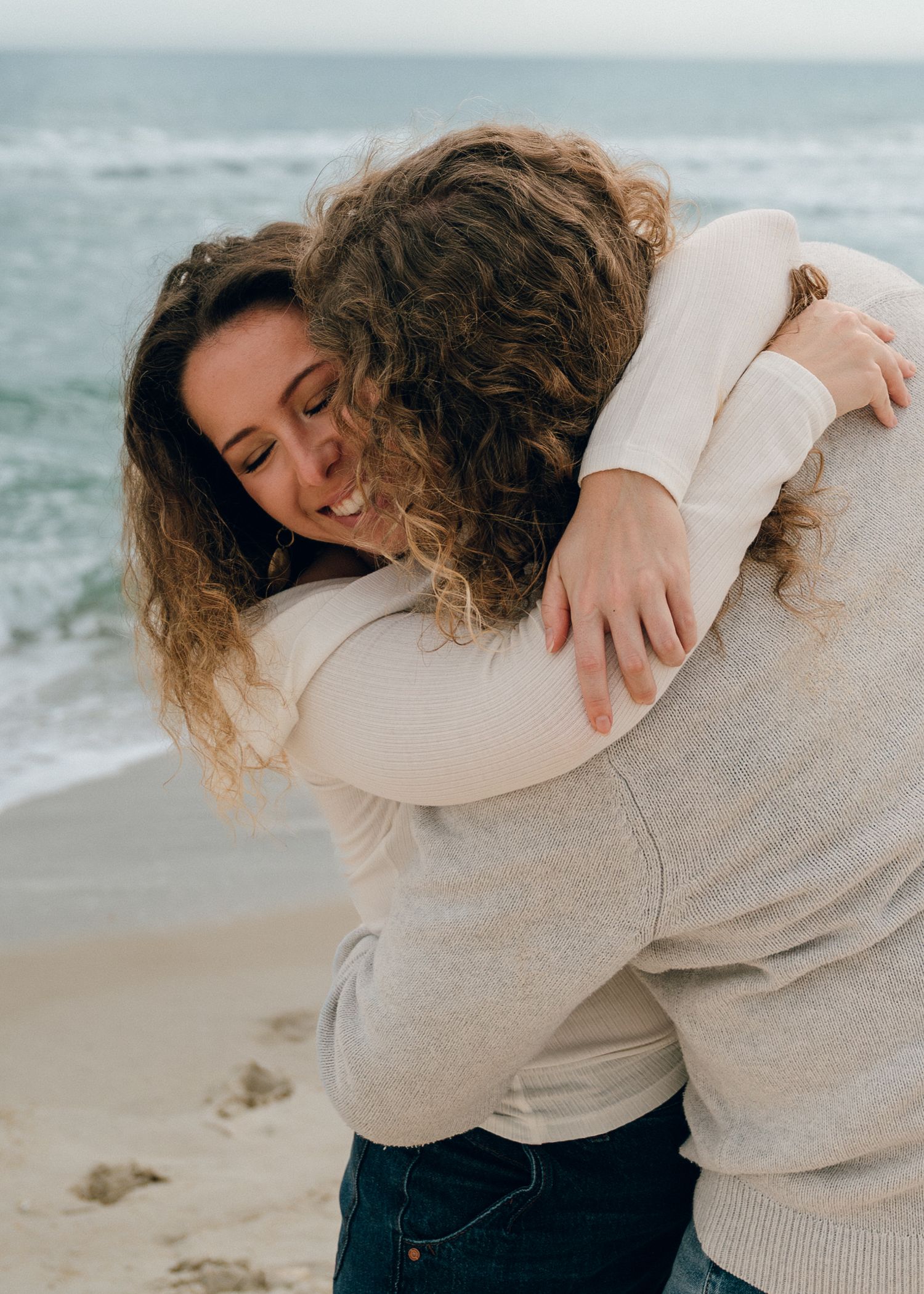 A couple embraces in a tender hug while standing on a beach with waves crashing in the background.