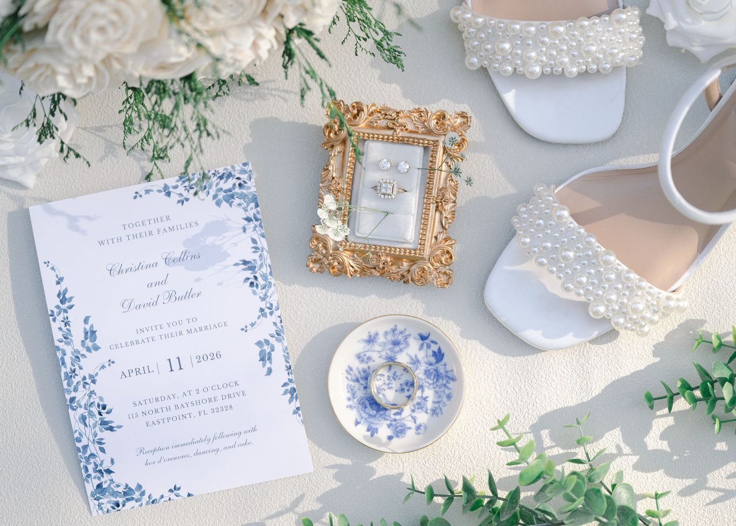 Blue floral wedding invitation flatlay with pearl heels, gold frame ring holder, and eucalyptus greenery on white background.