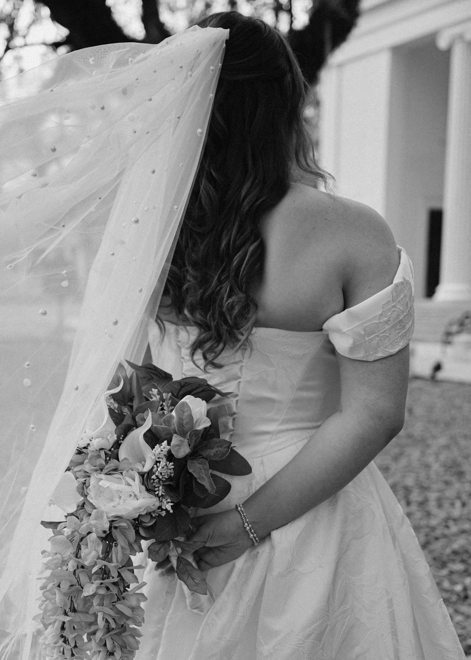 Black and white photo of a bride in an off-shoulder gown holding a floral bouquet, her veil flowing gracefully.