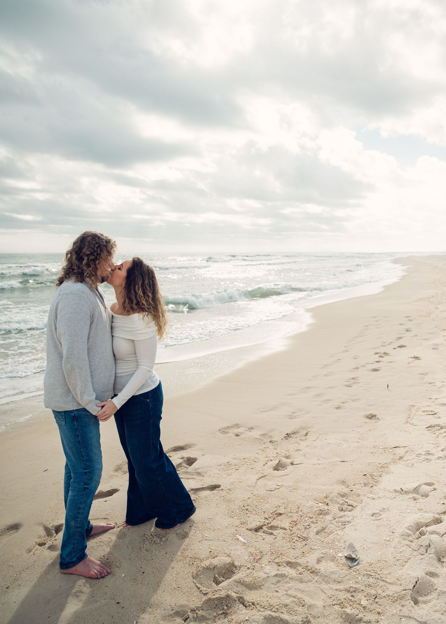 A couple shares a tender moment while walking along a misty beach shoreline on an overcast day.