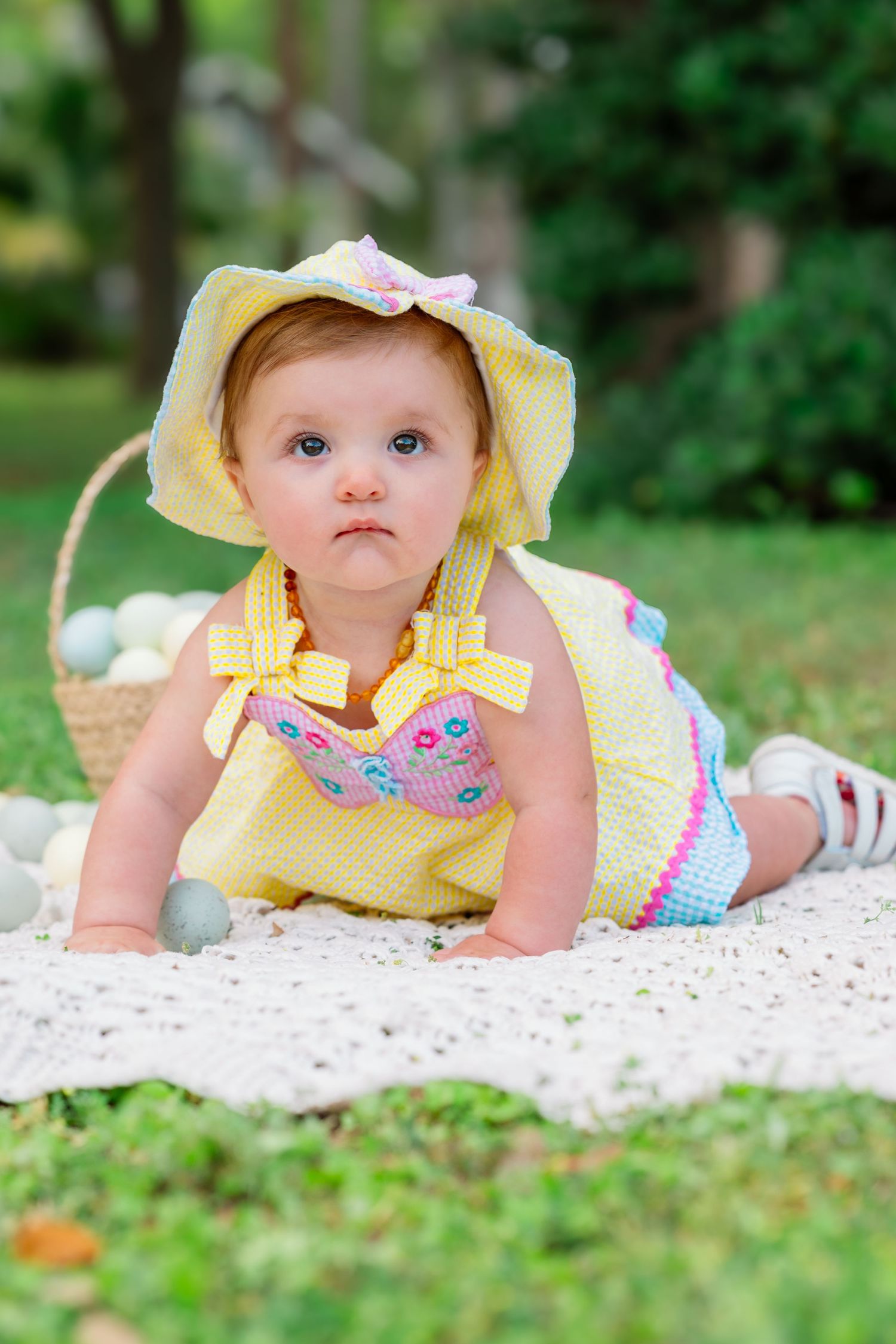Baby in yellow sunhat and floral outfit lies on white blanket outdoors.