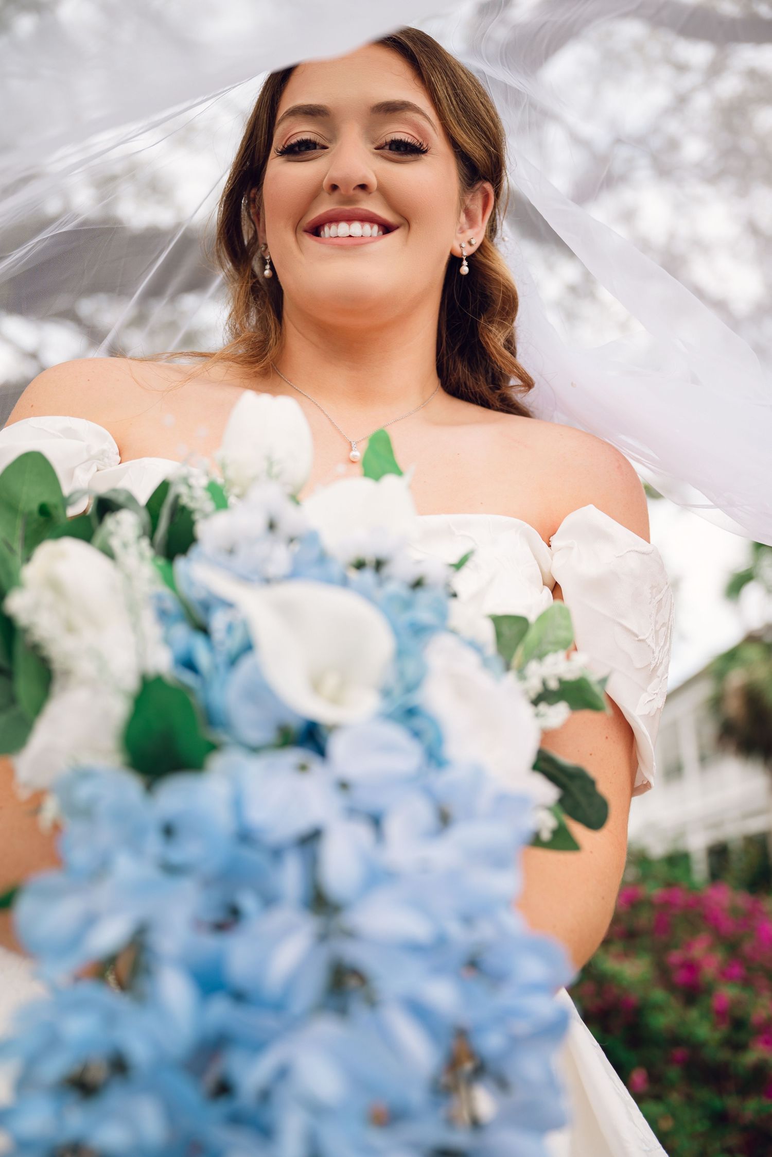 A bride in a white dress holds a beautiful bouquet featuring light blue hydrangeas and white flowers outdoors.