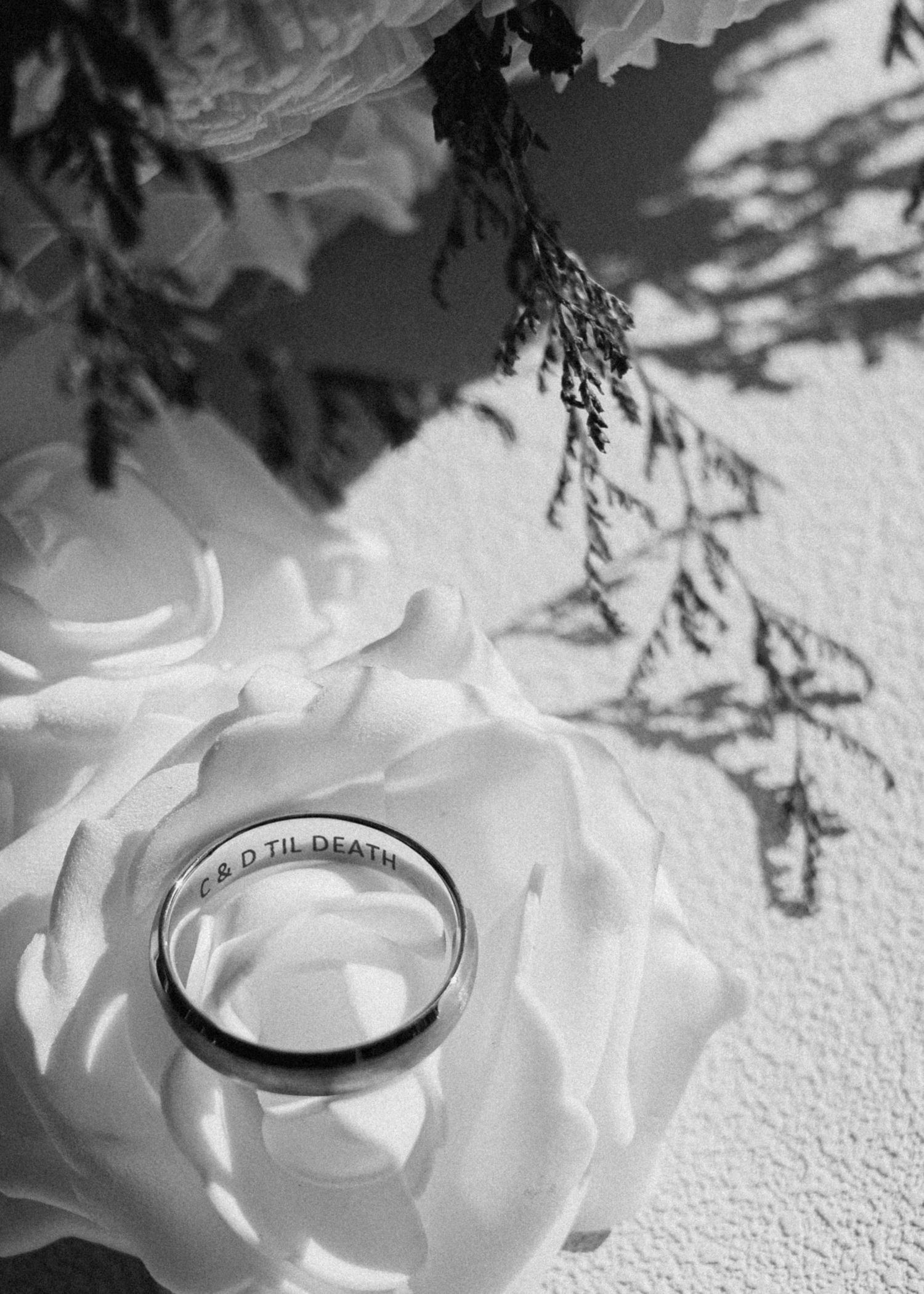 Black and white photo of a wedding band engraved with 'TIL DEATH' resting on a white rose with floral shadows.