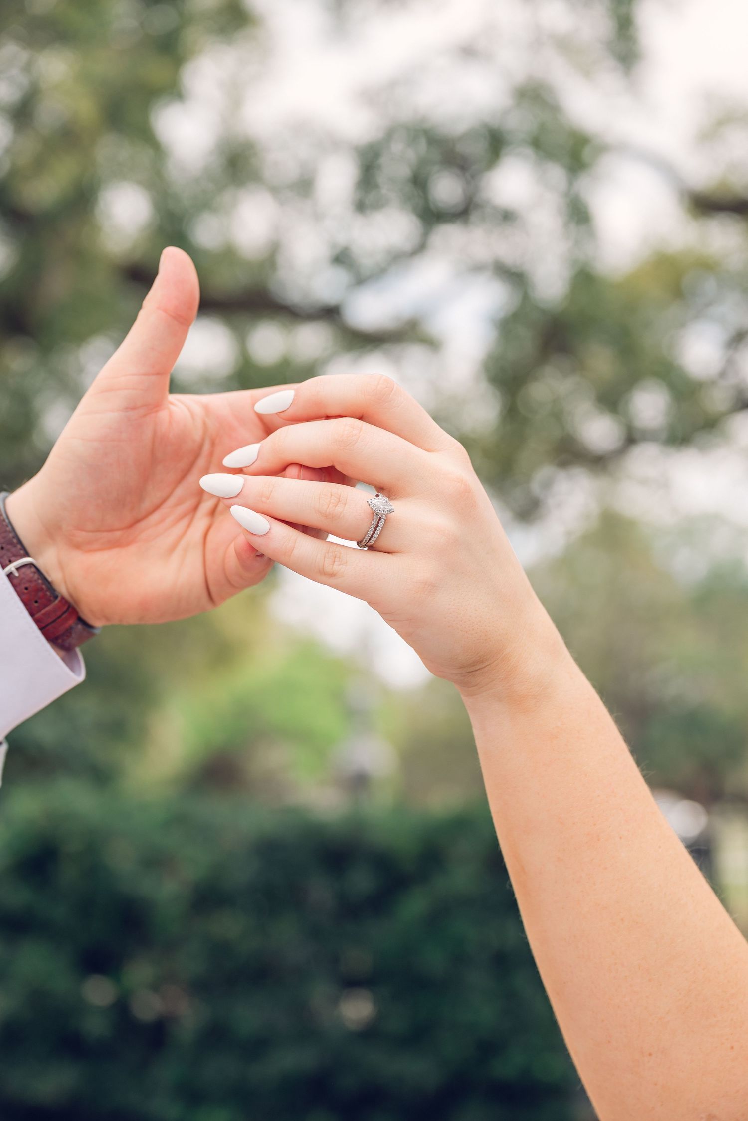 Two hands gently touch as one displays a wedding ring against a soft, blurred green outdoor background.