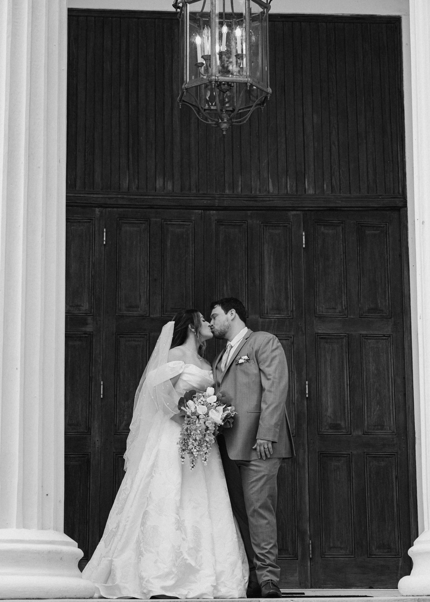 Black and white photo of a bride and groom sharing a kiss outside large wooden doors with a vintage lantern above.