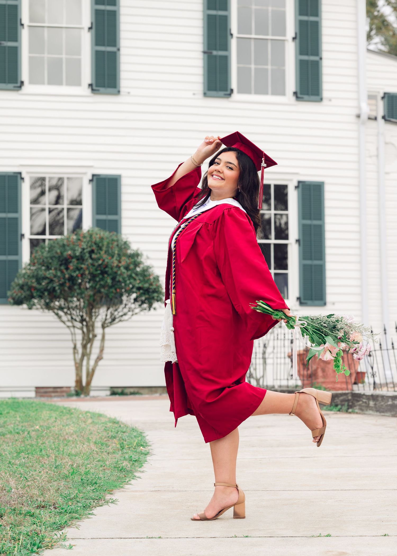 A graduate in a red cap and gown poses in front of a white colonial style house with green shutters in three dynamic poses.
