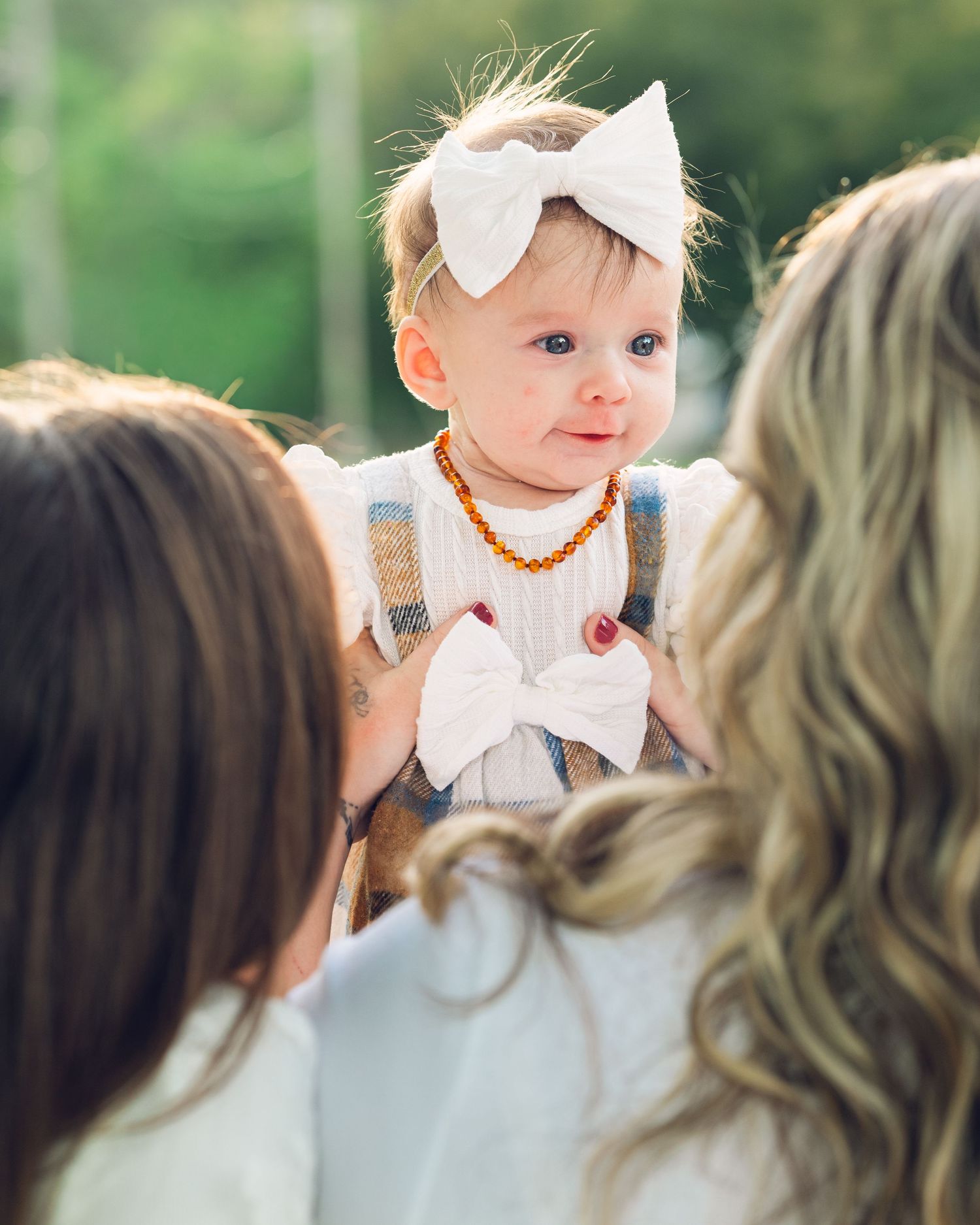 A baby wearing a white bow and amber necklace is held up during an outdoor gathering.
