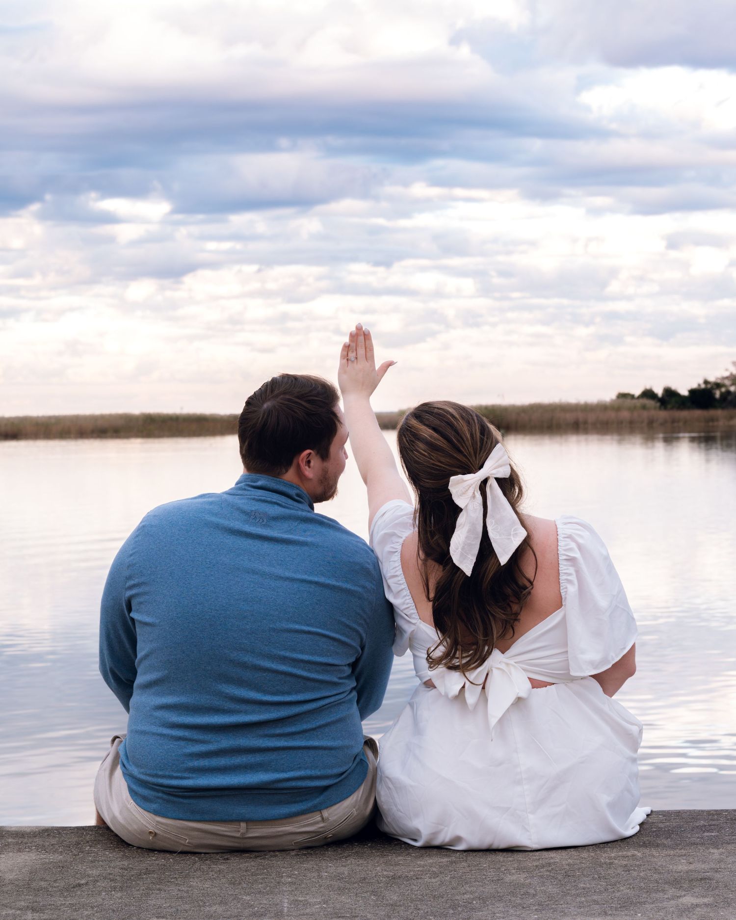 A couple sitting on a dock overlooking a serene lake with cloudy skies.