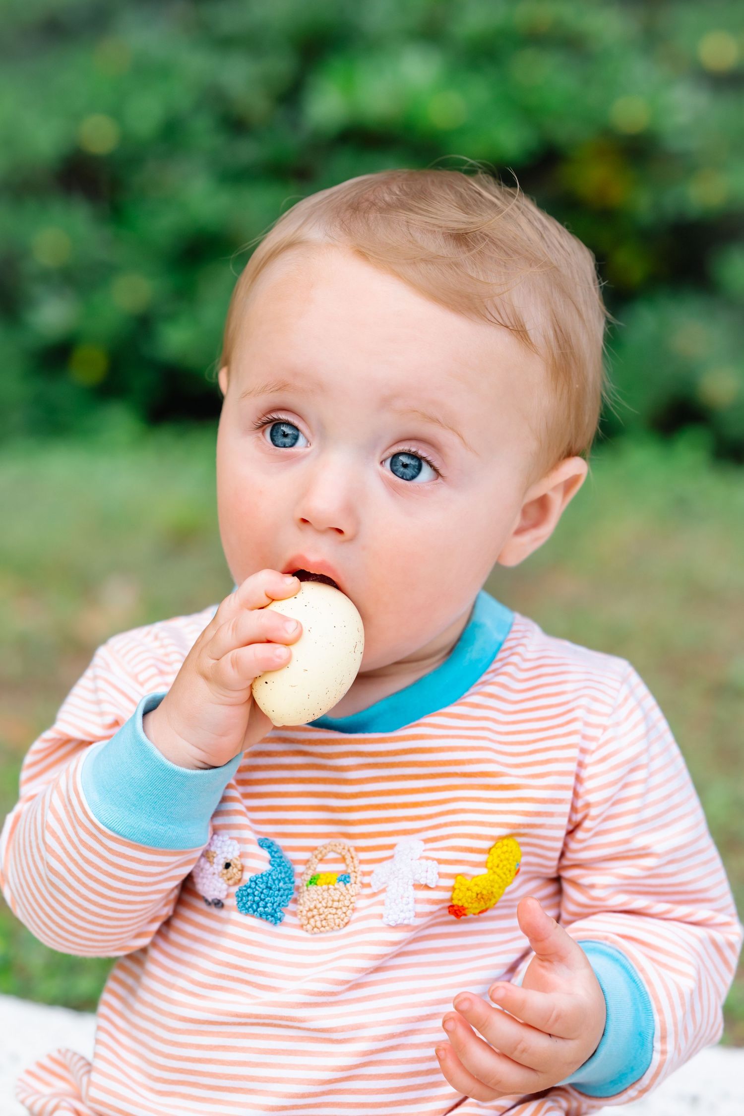 Baby in striped outfit with blue bib eating snacks outdoors.