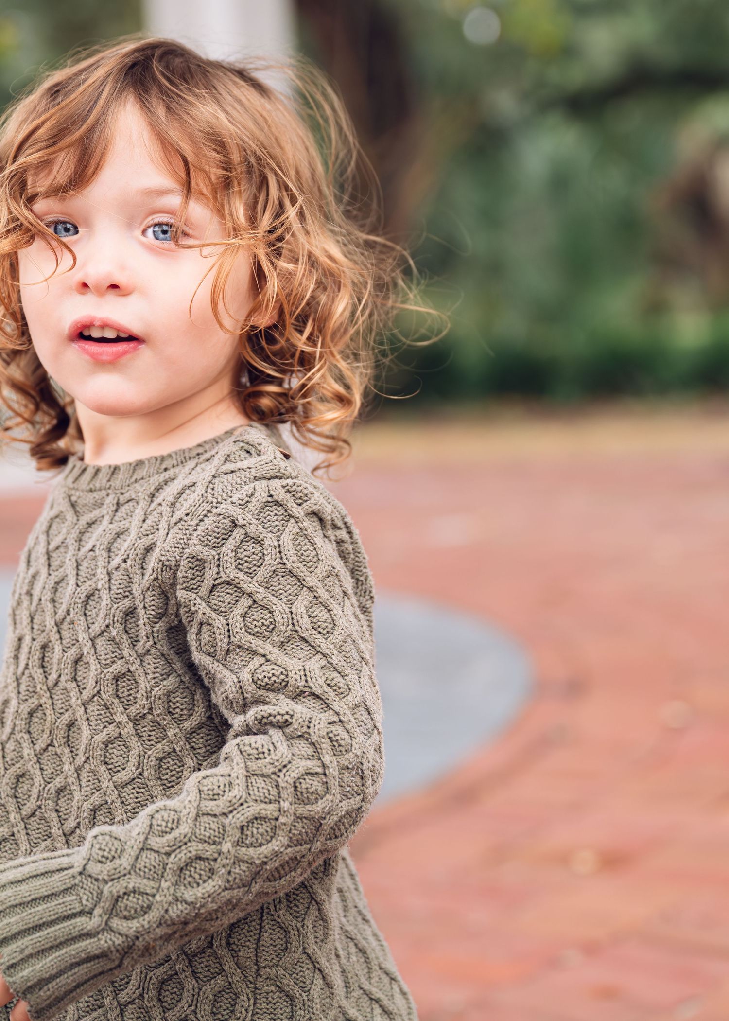Child in grey knit sweater poses outdoors against blurred natural background.