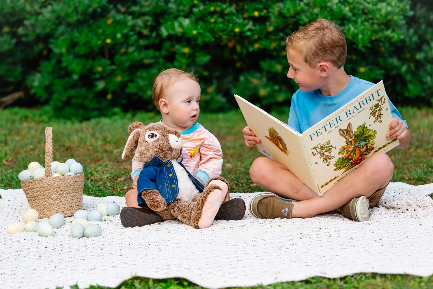 Children and adults read books together on white blanket outdoors.