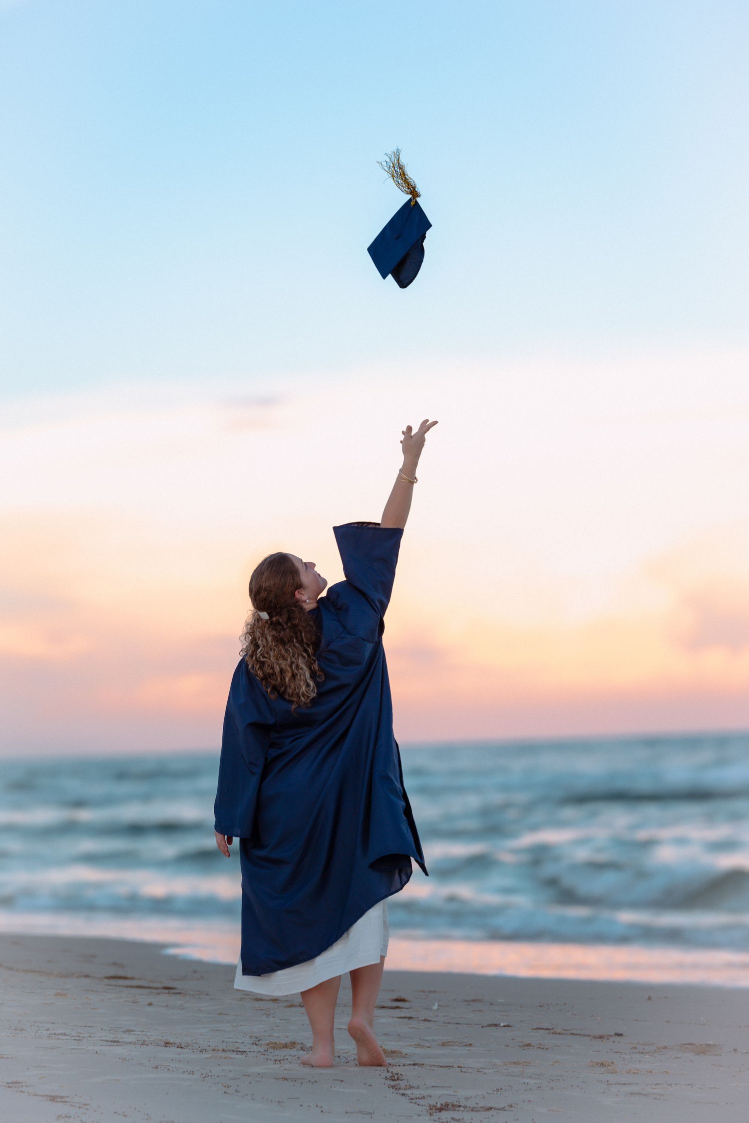 Graduate throwing cap on beach at sunset in celebration against ocean waves.