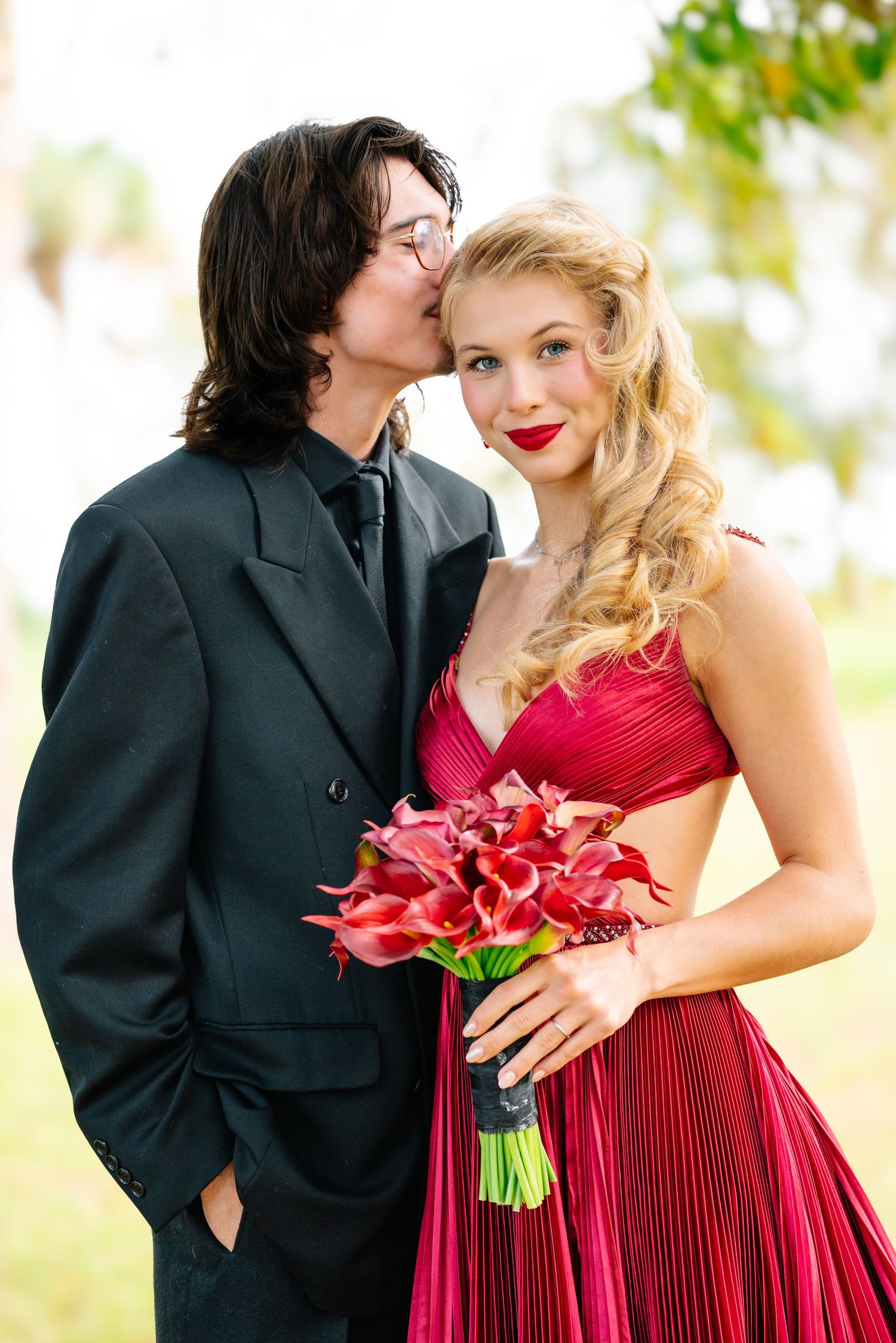 Romantic couple poses in formal wear with red flowers against a bright outdoor setting.