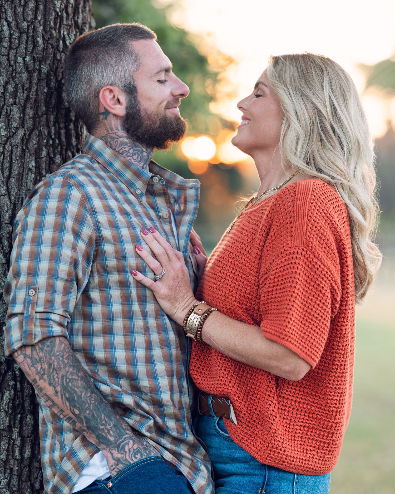 Engagement photos showing couple in casual attire leaning against large tree.