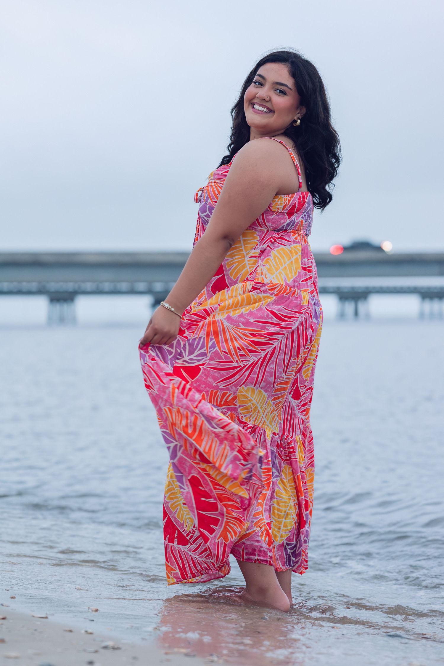 Someone wearing a vibrant pink and orange tropical print maxi dress poses joyfully at the beach during sunset.