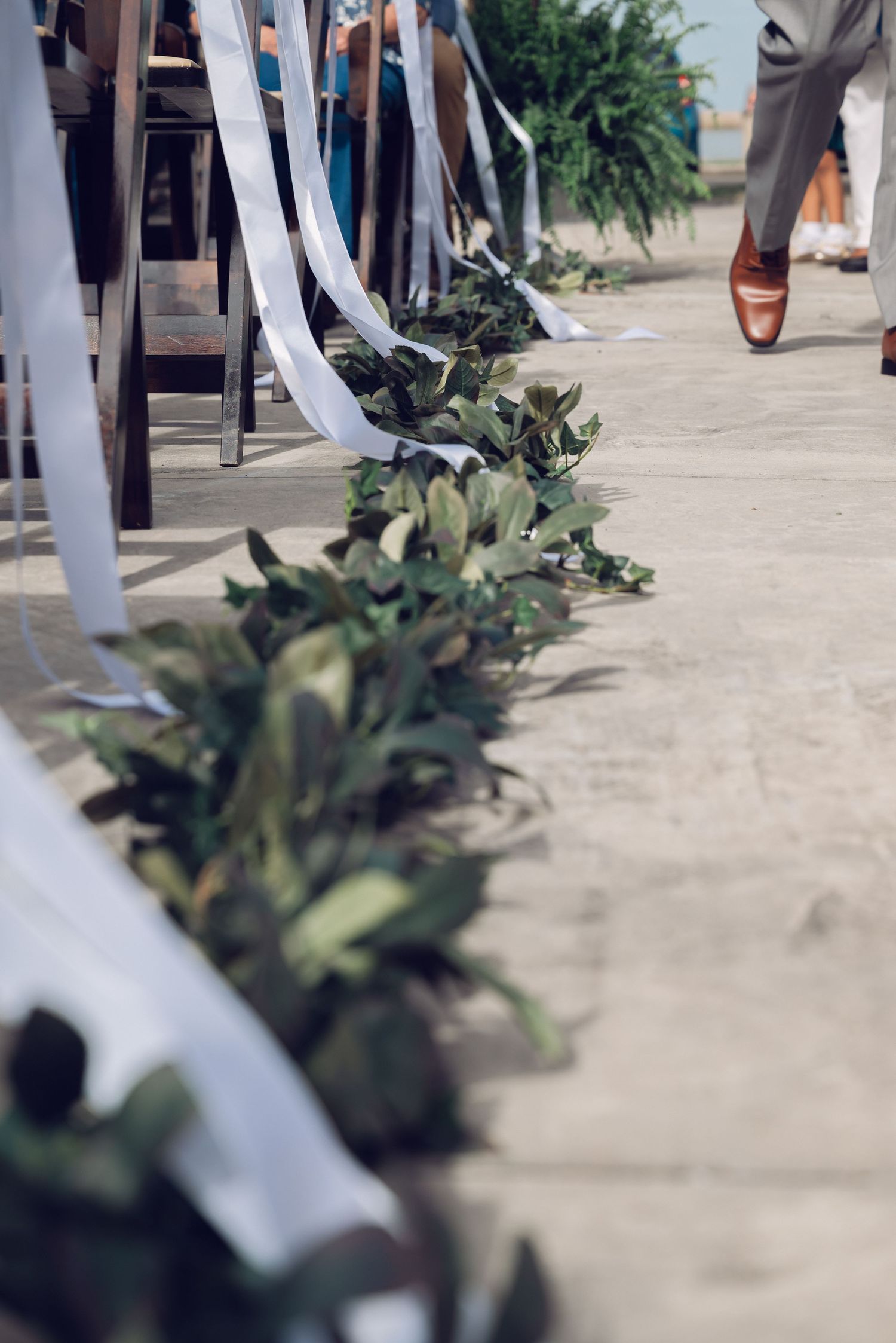 Green leaf garland lines a wedding ceremony aisle with white ribbon chair decor and a guest walking in brown dress shoes.