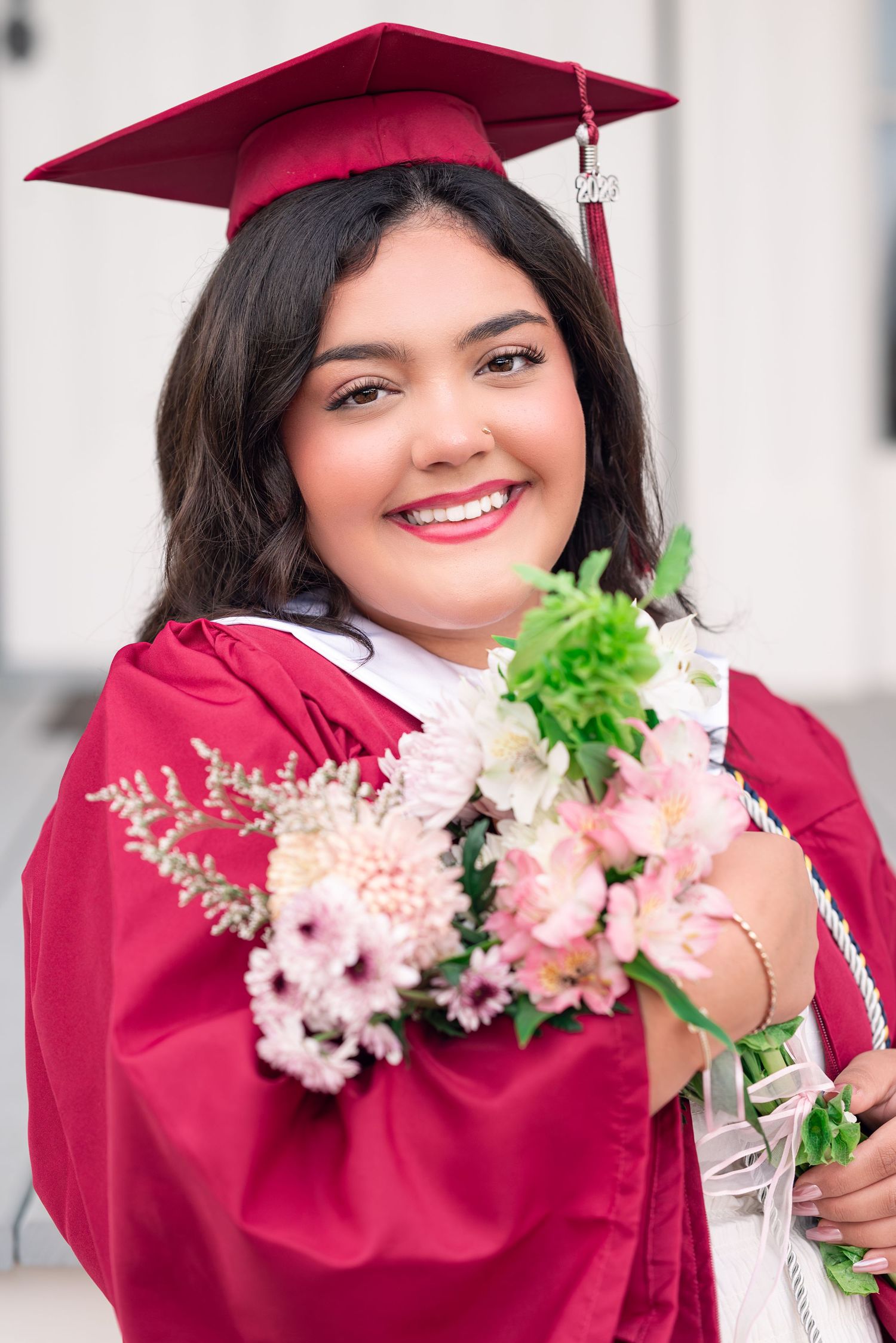 A graduate in a burgundy cap and gown holds a bouquet of pink and white flowers with greenery.