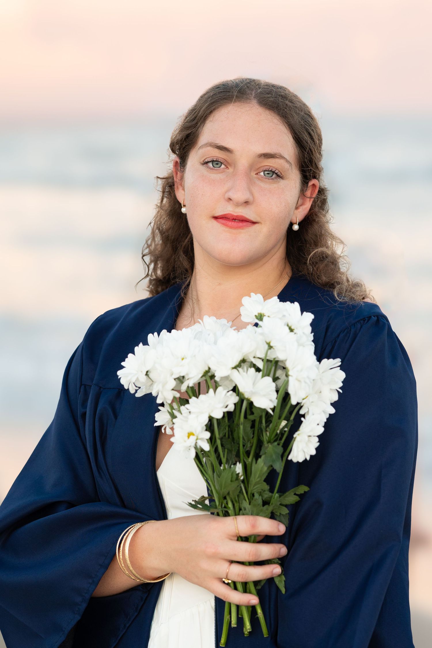 Graduate in navy blue gown holding white flowers during beach photo session.