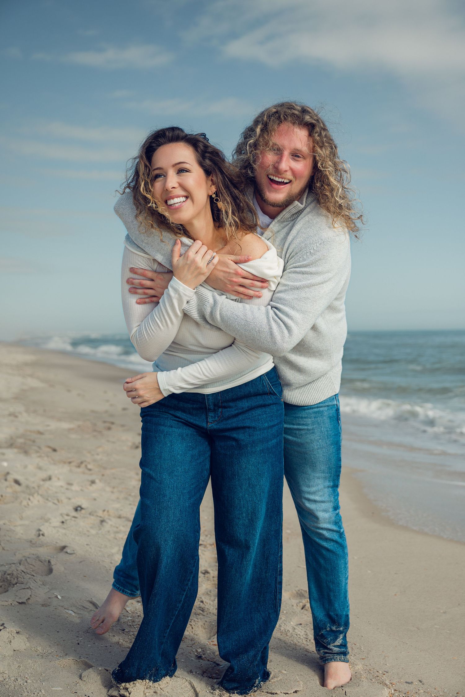 Couple in matching white sweaters and jeans embrace joyfully on a sunny beach with ocean waves in the background.
