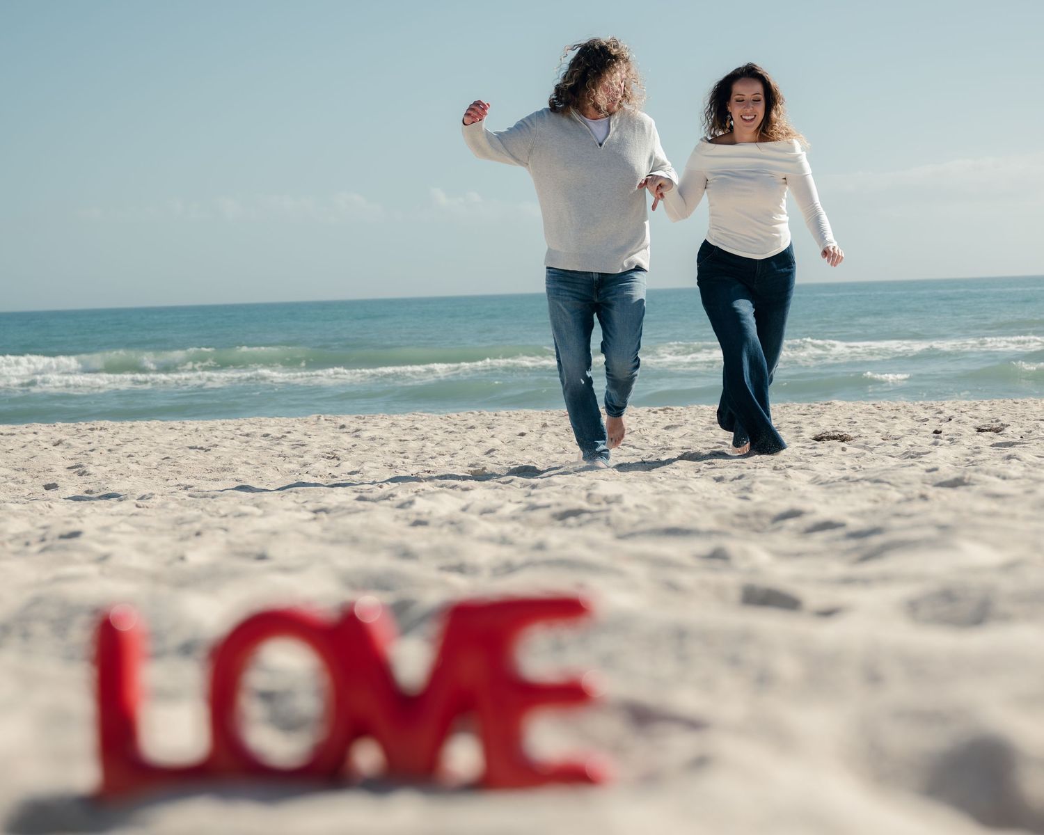Couple walking hand in hand on a sandy beach with ocean waves in the background and red love text on sand.
