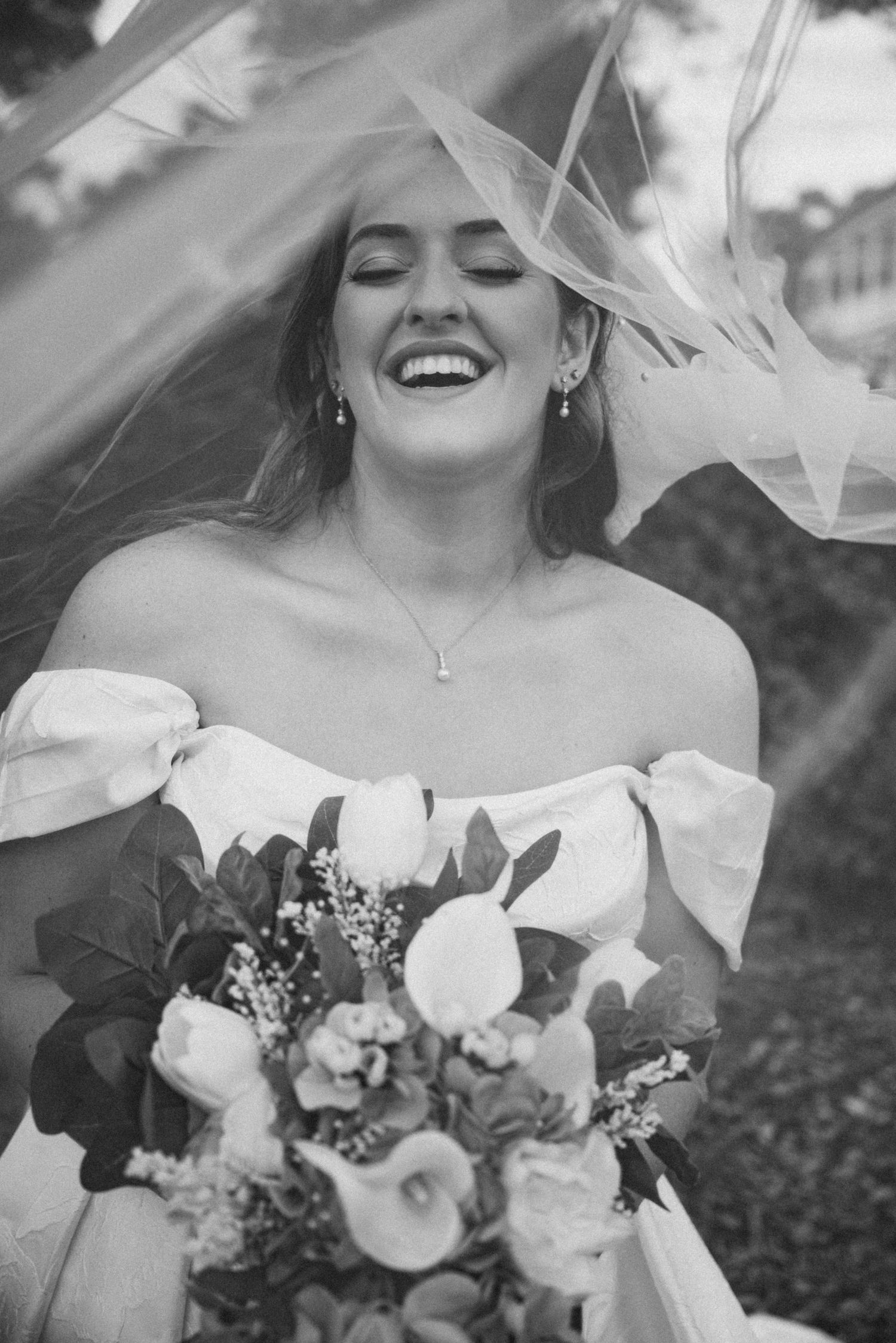 A joyful bride in a white off-shoulder gown laughs while holding a bouquet of white flowers under a flowing veil.