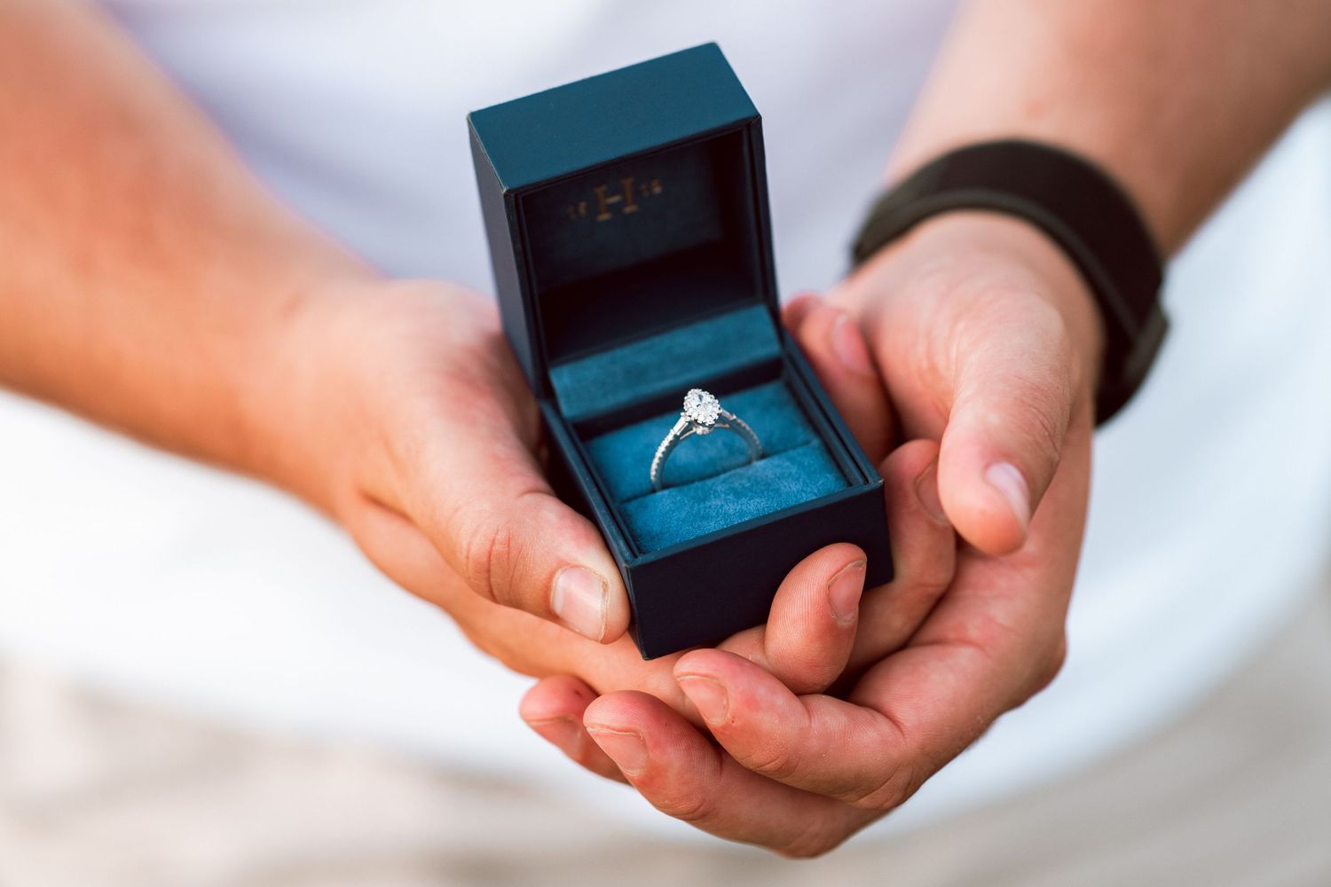 Hands holding blue engagement ring box against white background.