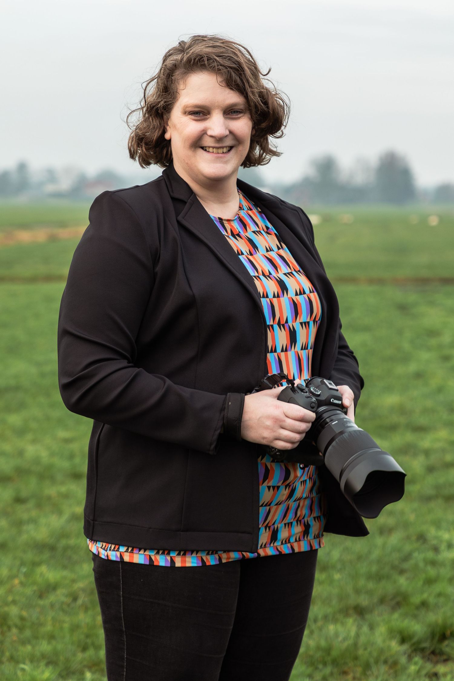 Smiling woman holding a professional camera outdoors, wearing a colorful top and black blazer, standing in a green field.
