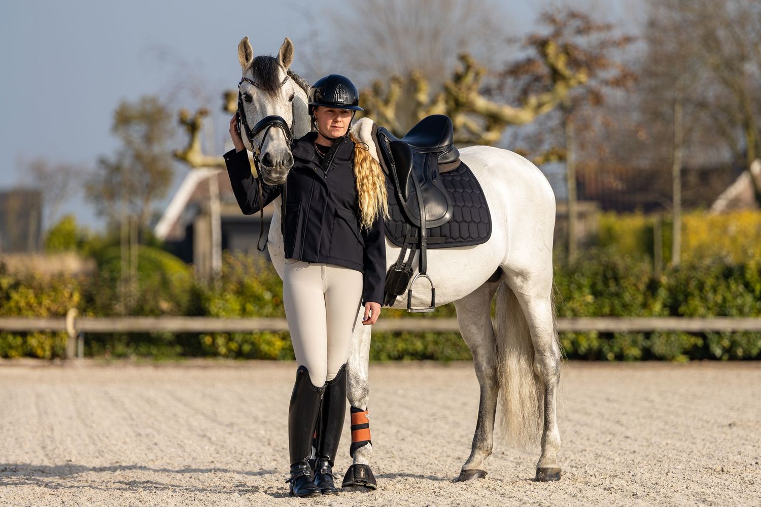 Equestrian rider in black jacket and helmet standing beside a white horse on a sandy arena outdoors.