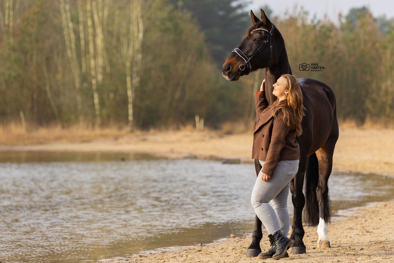 Woman in brown jacket standing beside a dark bay horse on a sandy riverbank with autumn trees in background.