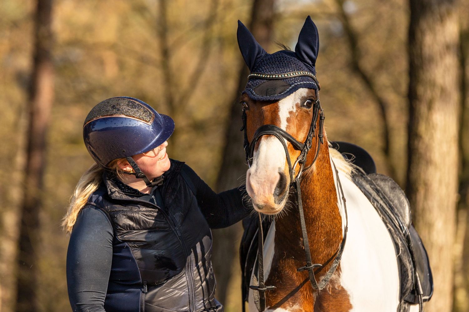 A female equestrian in a navy helmet stands beside a brown and white horse wearing a fly veil in an autumn forest.