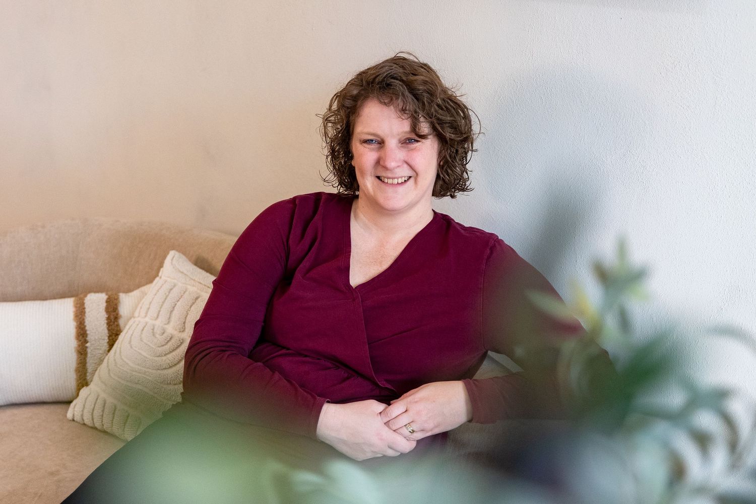 A smiling woman in a burgundy top sits relaxed on a cozy sofa with decorative pillows in a bright, modern room.