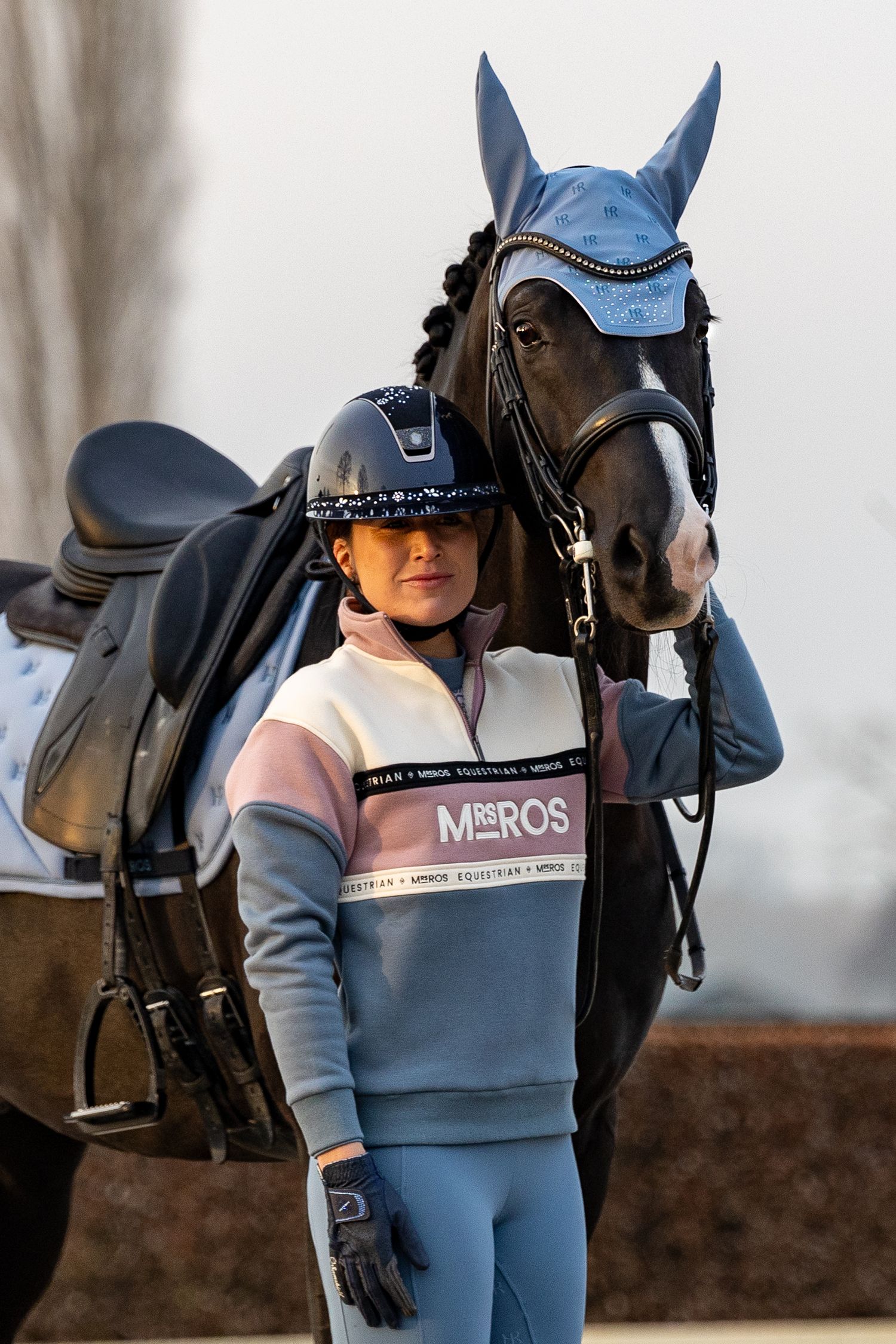 Young equestrian rider in MosRos sweatshirt and helmet standing beside a dark bay horse with blue ear bonnet.