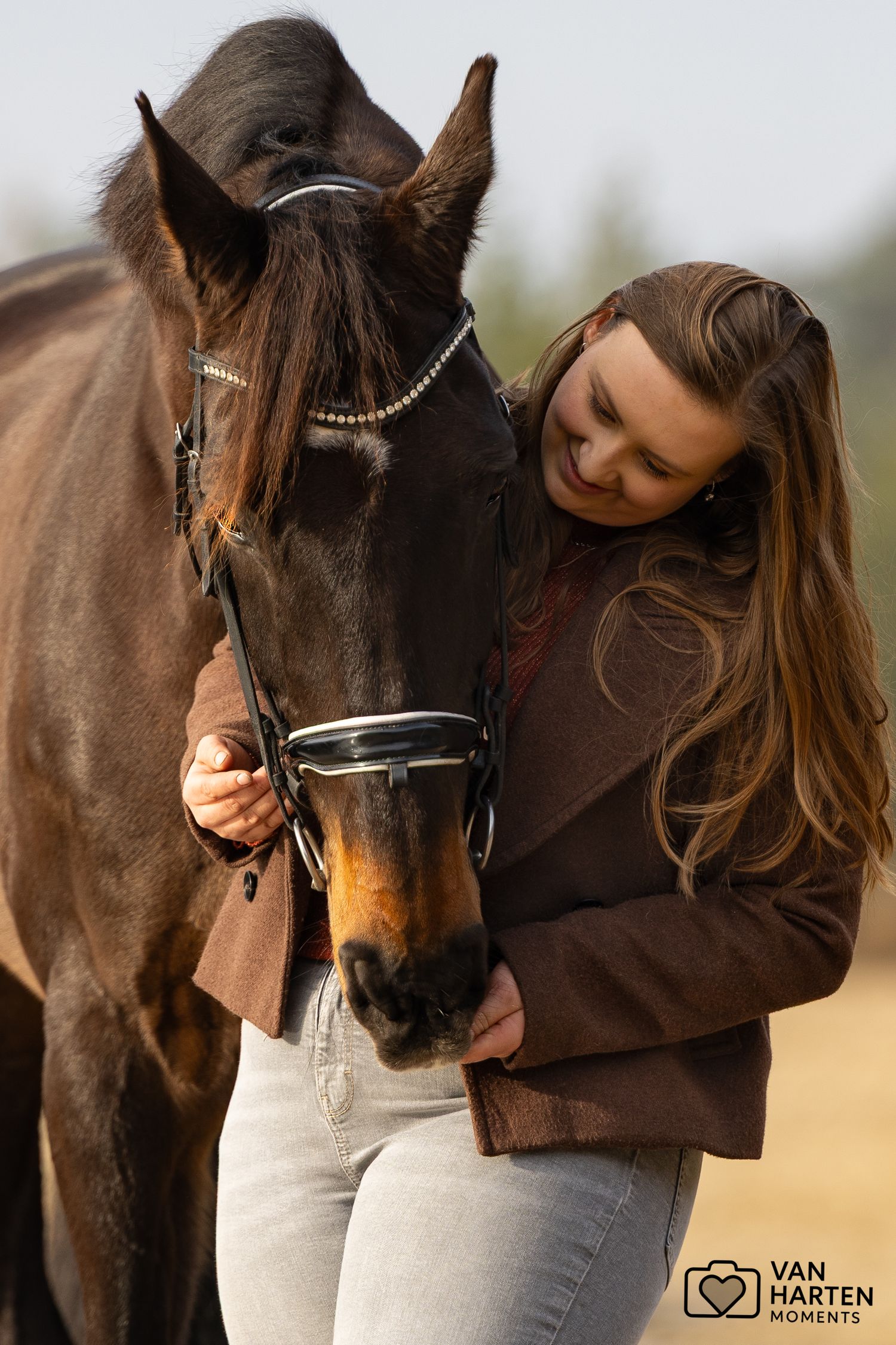 A young woman with long hair tenderly embraces a dark brown horse in an open field.