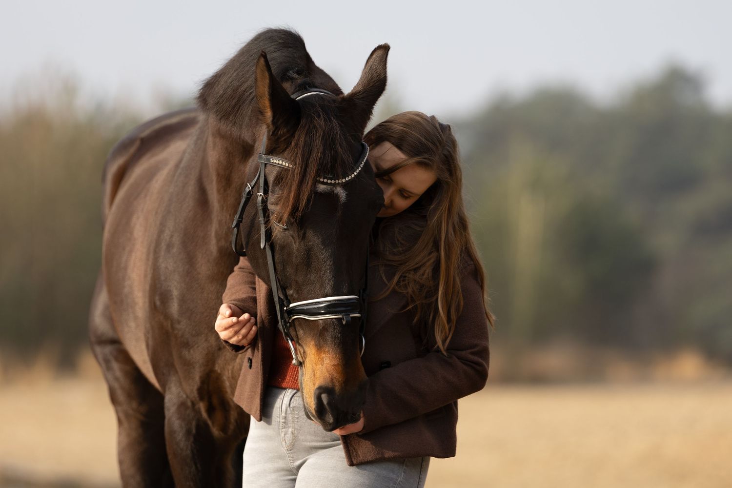 Young woman in brown jacket tenderly embracing a dark brown horse in an open field.