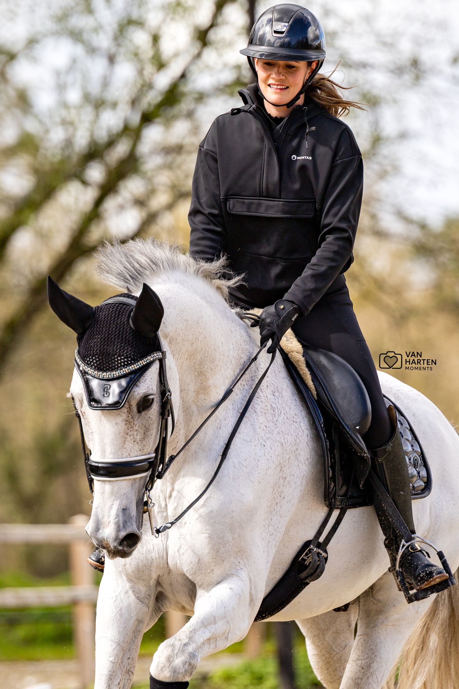 Smiling woman in black riding gear on a white horse during an outdoor dressage session.