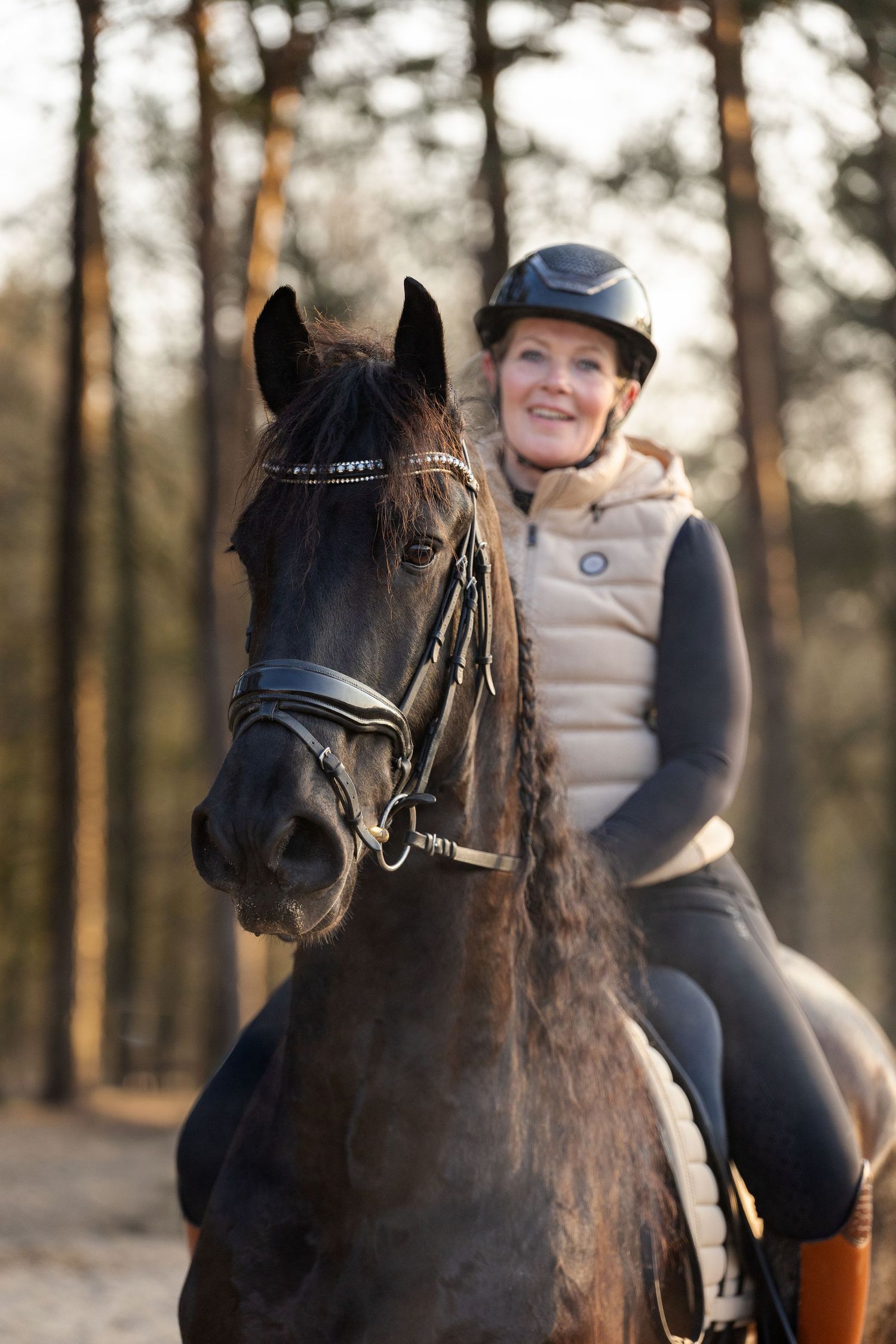 A smiling woman in a riding helmet sits atop a dark horse in a sunlit forest setting.