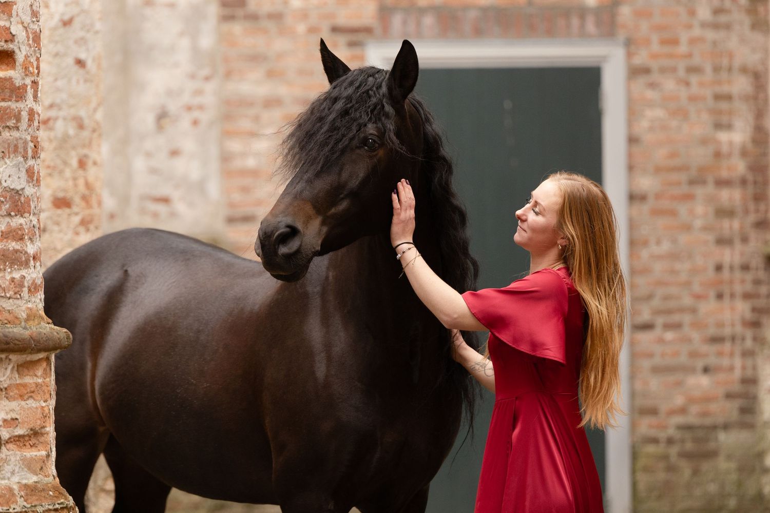 A woman in a red dress gently touches a dark brown horse near a rustic brick building.