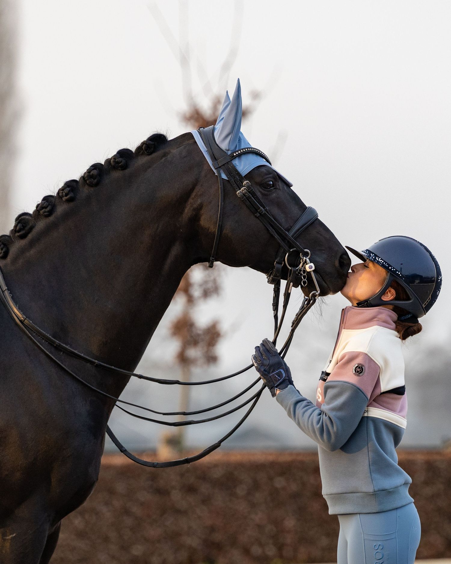 A rider in a helmet shares a tender moment kissing her dark black horse wearing a blue ear bonnet and bridle.