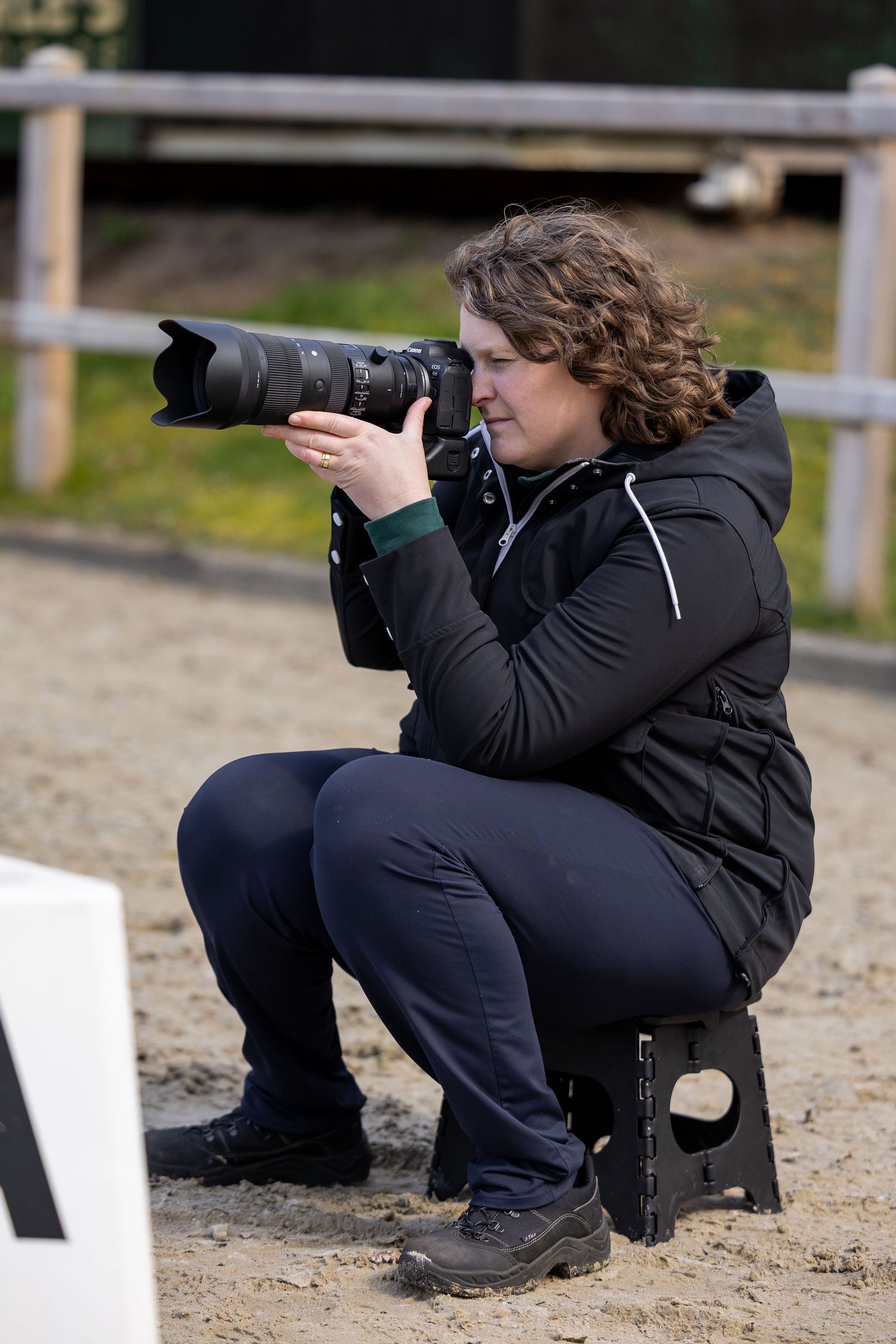 A photographer with curly hair crouches on a stool, aiming a telephoto lens camera outdoors near a sandy arena.
