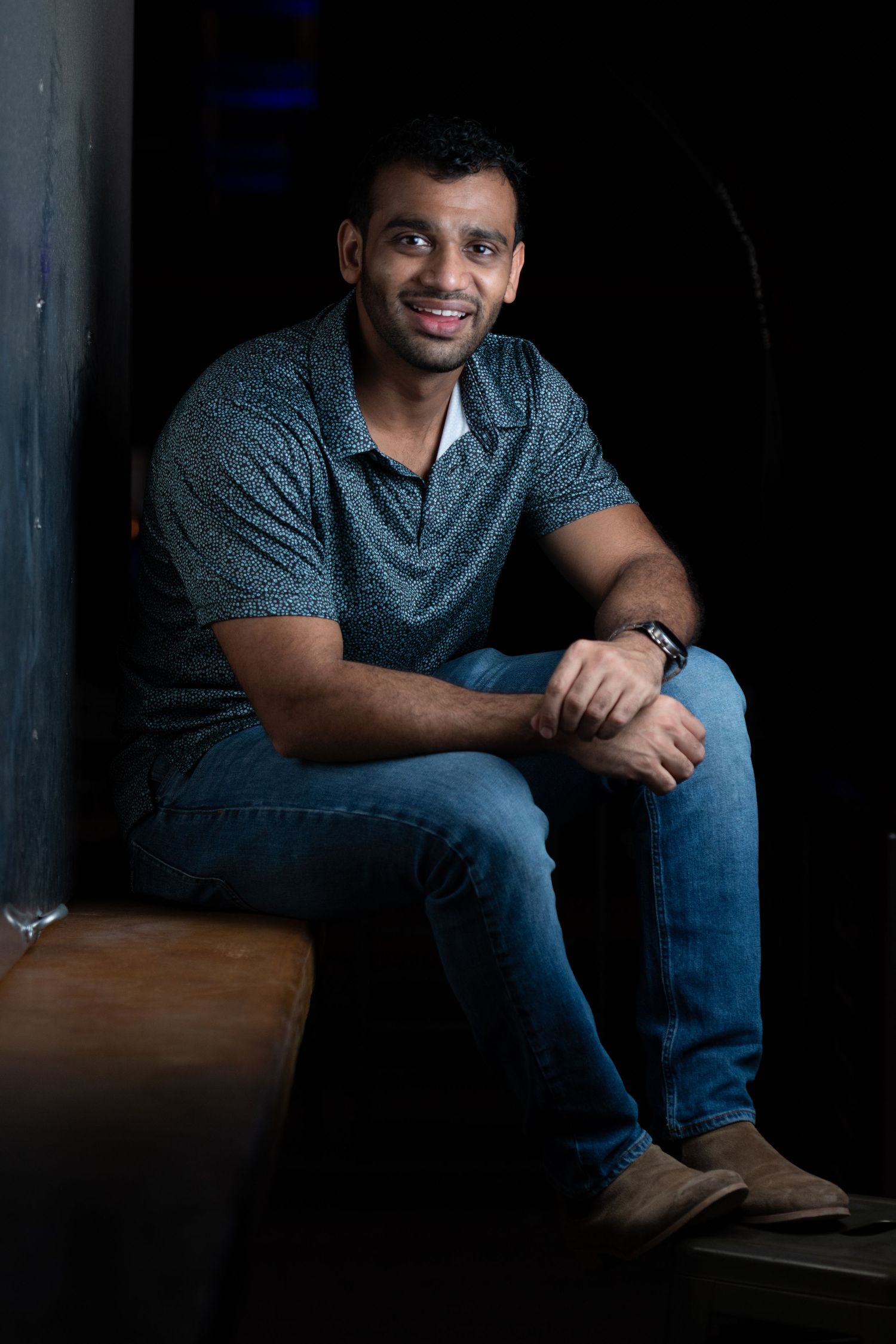 A person in jeans and a gray polo shirt sits casually on a bench in a dimly lit setting.